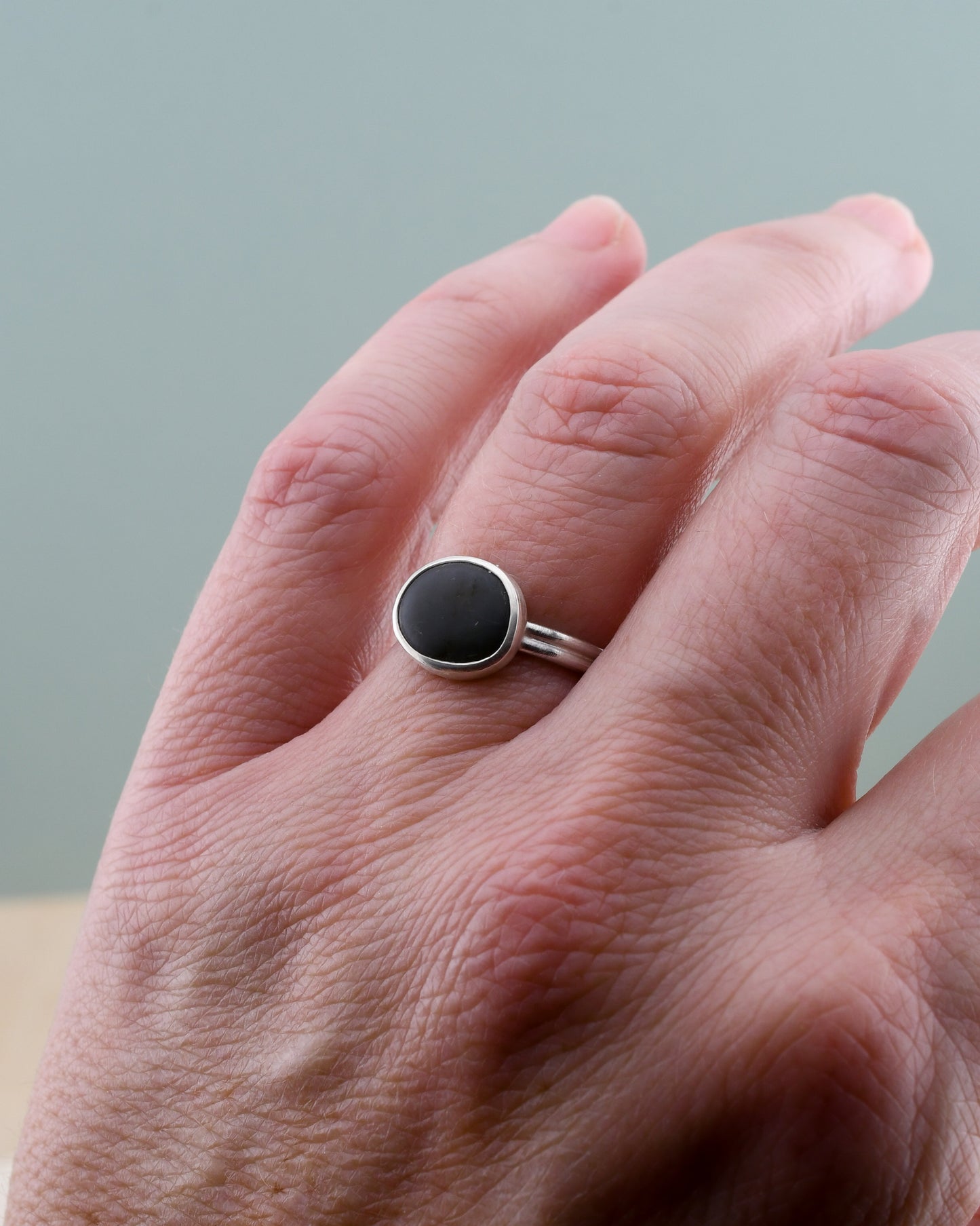Hand wearing a silver ring with a dark gray stone on a neutral background