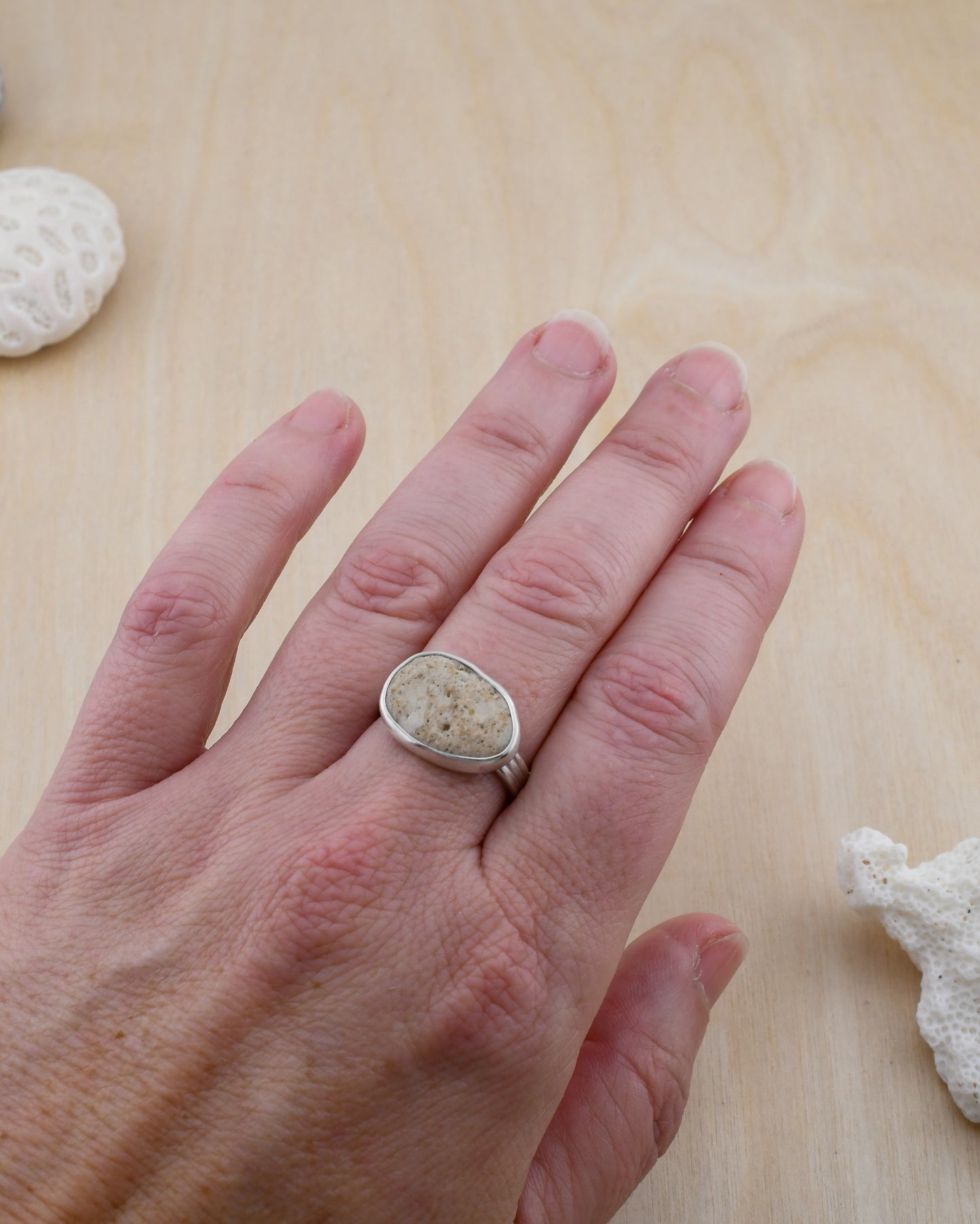 Hand wearing a ring with a textured stone design on a wooden surface with coral.