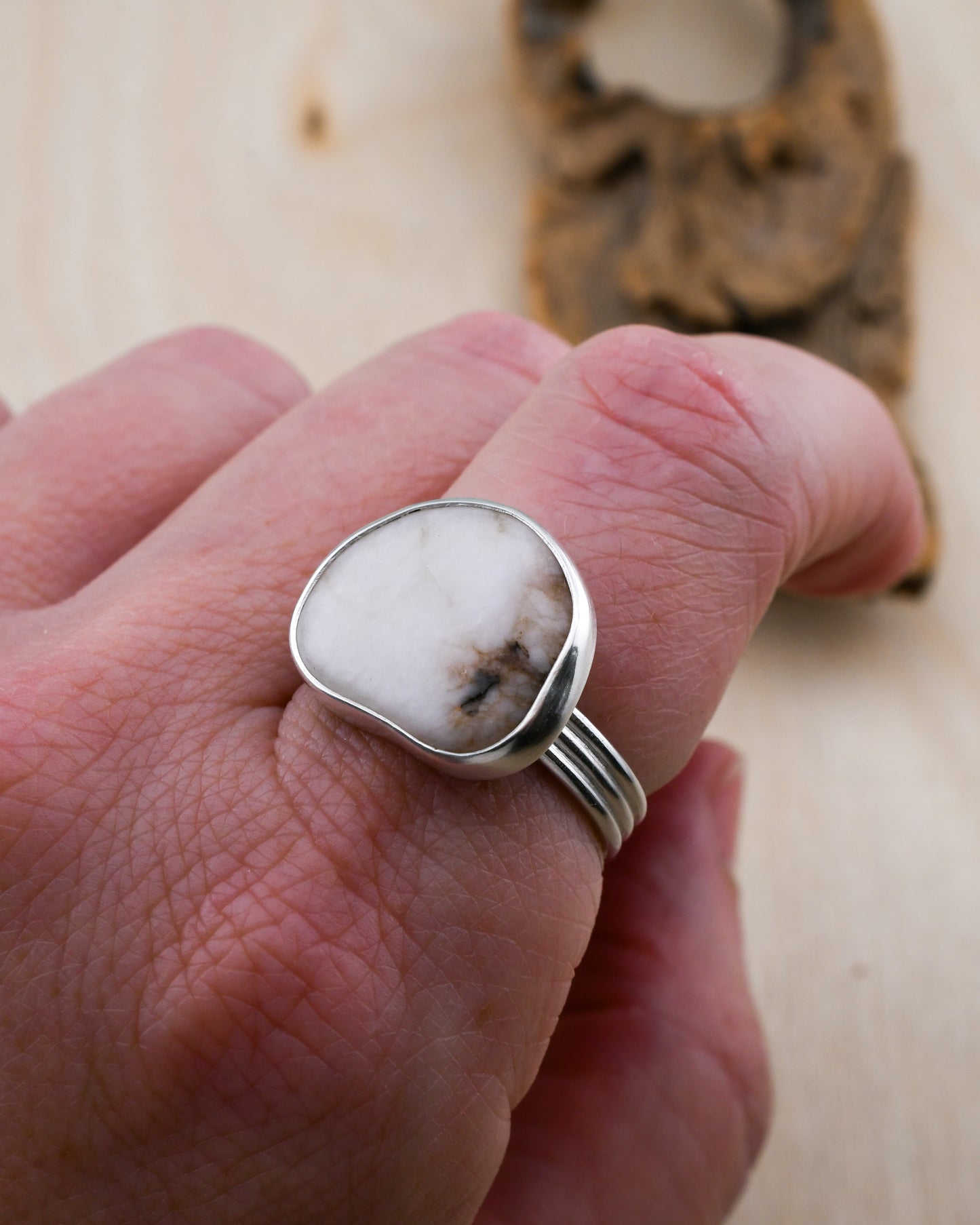 Hand wearing a silver ring with a large, irregularly shaped white beach stone on a wooden surface.