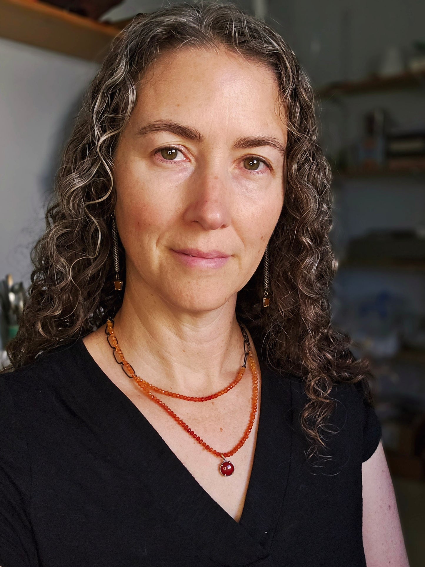 Woman with curly hair wearing a black top and orange beaded necklace indoors.