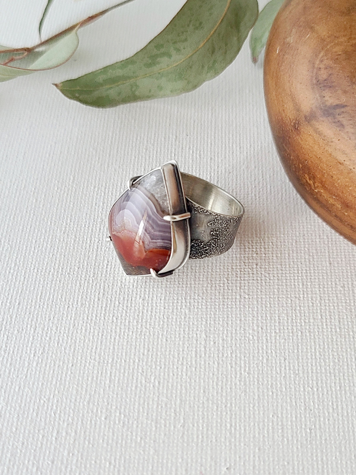 Red and purple Laguna lace agate ring with a wide textured band shown from above on a white linen surface with leaves and a wooden bowl
