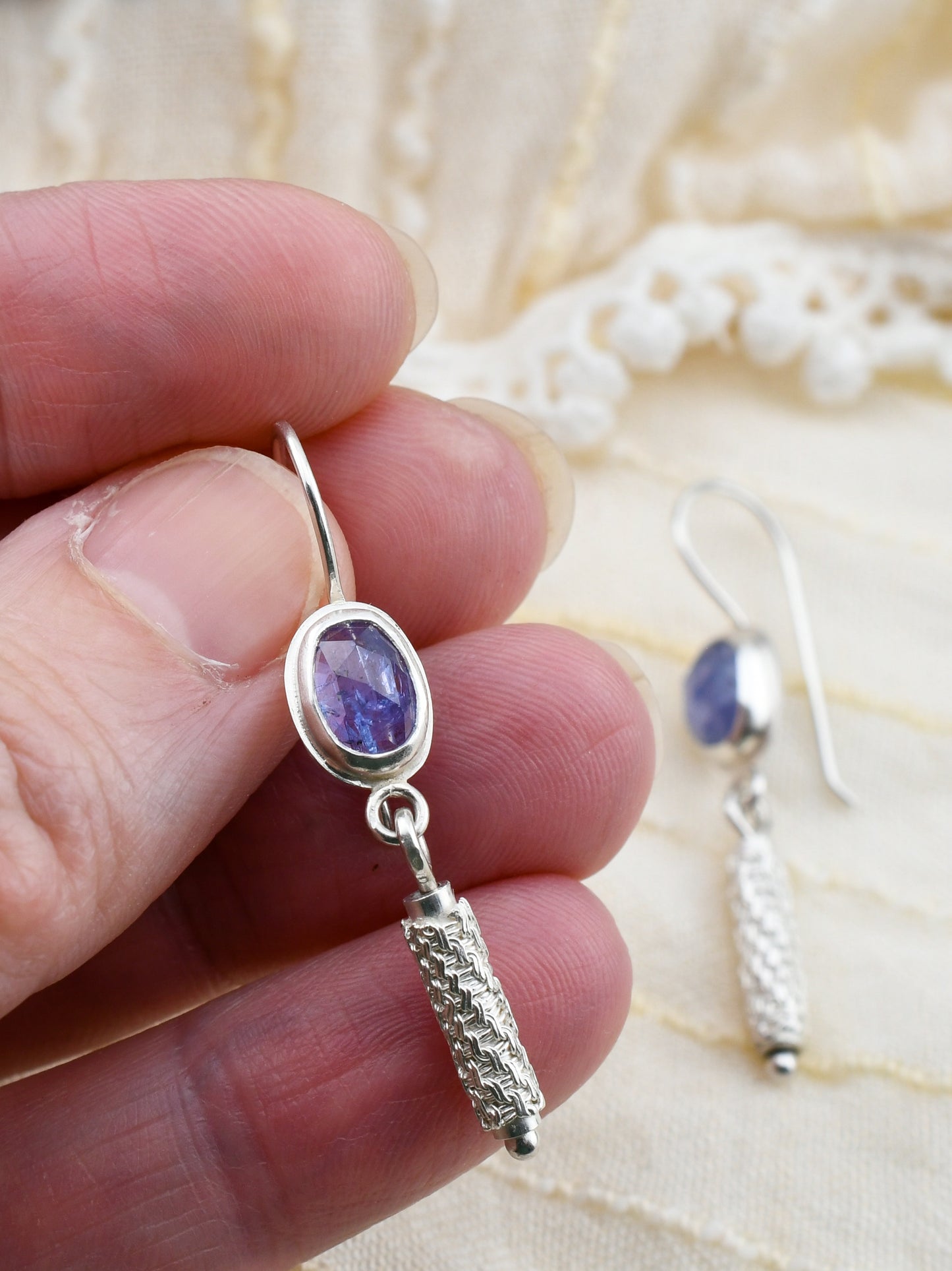 Silver earring with purple gemstone held between fingers on a light fabric background