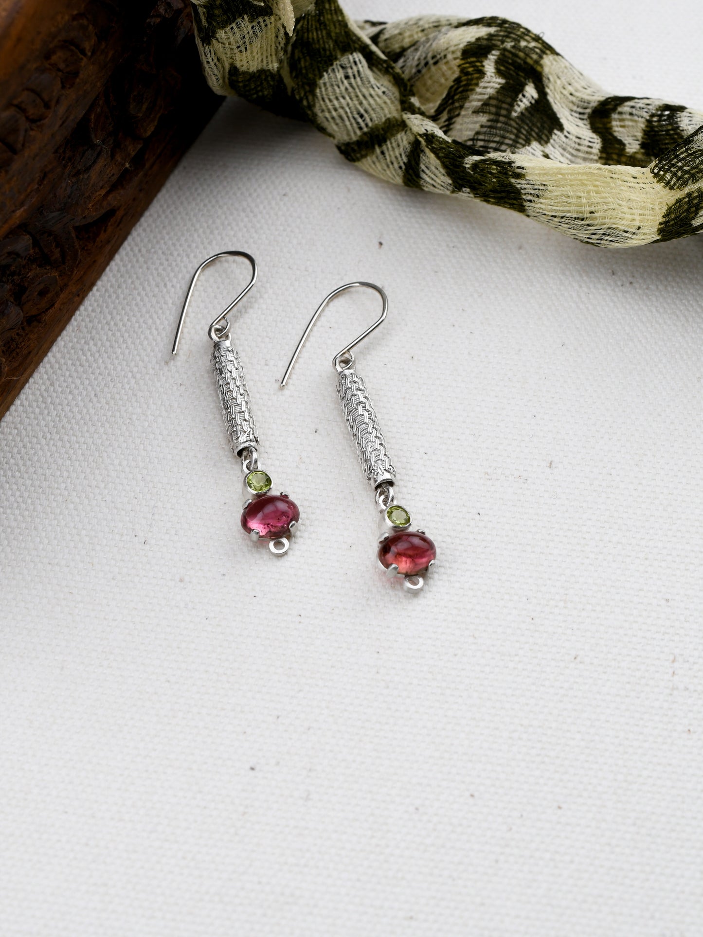 Silver earrings with pink and green stones on a white surface with a patterned fabric background.
