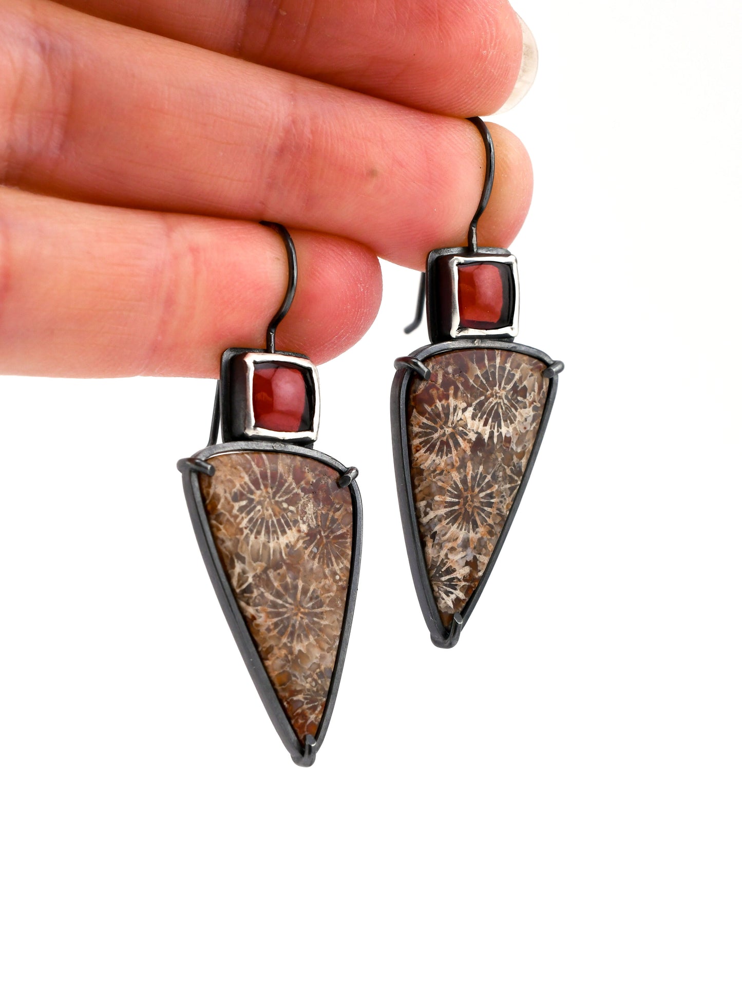 Triangular earrings with brown and red stones held by a hand on a white background