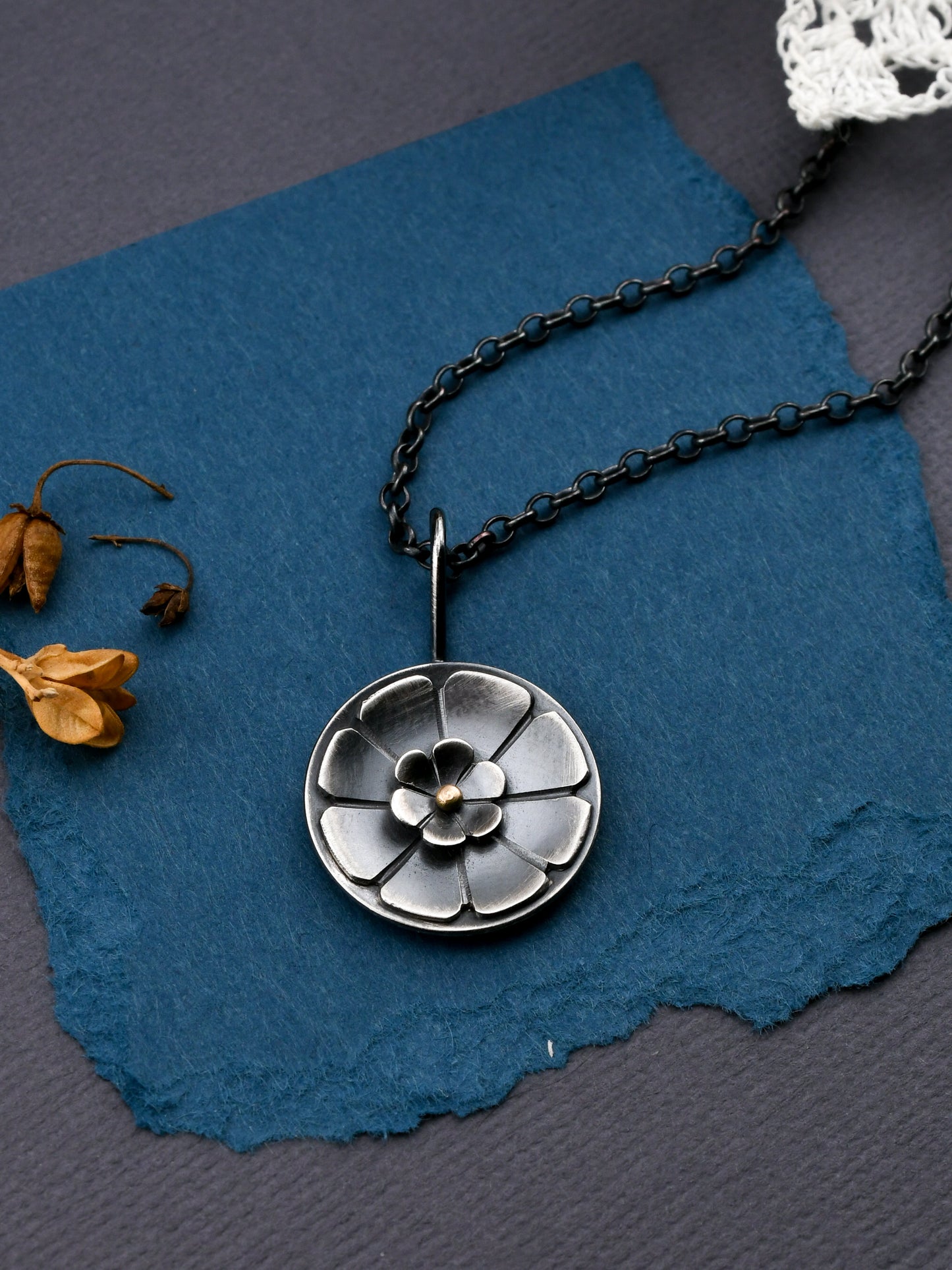 Silver floral pendant necklace on a blue textured surface with dried flowers.
