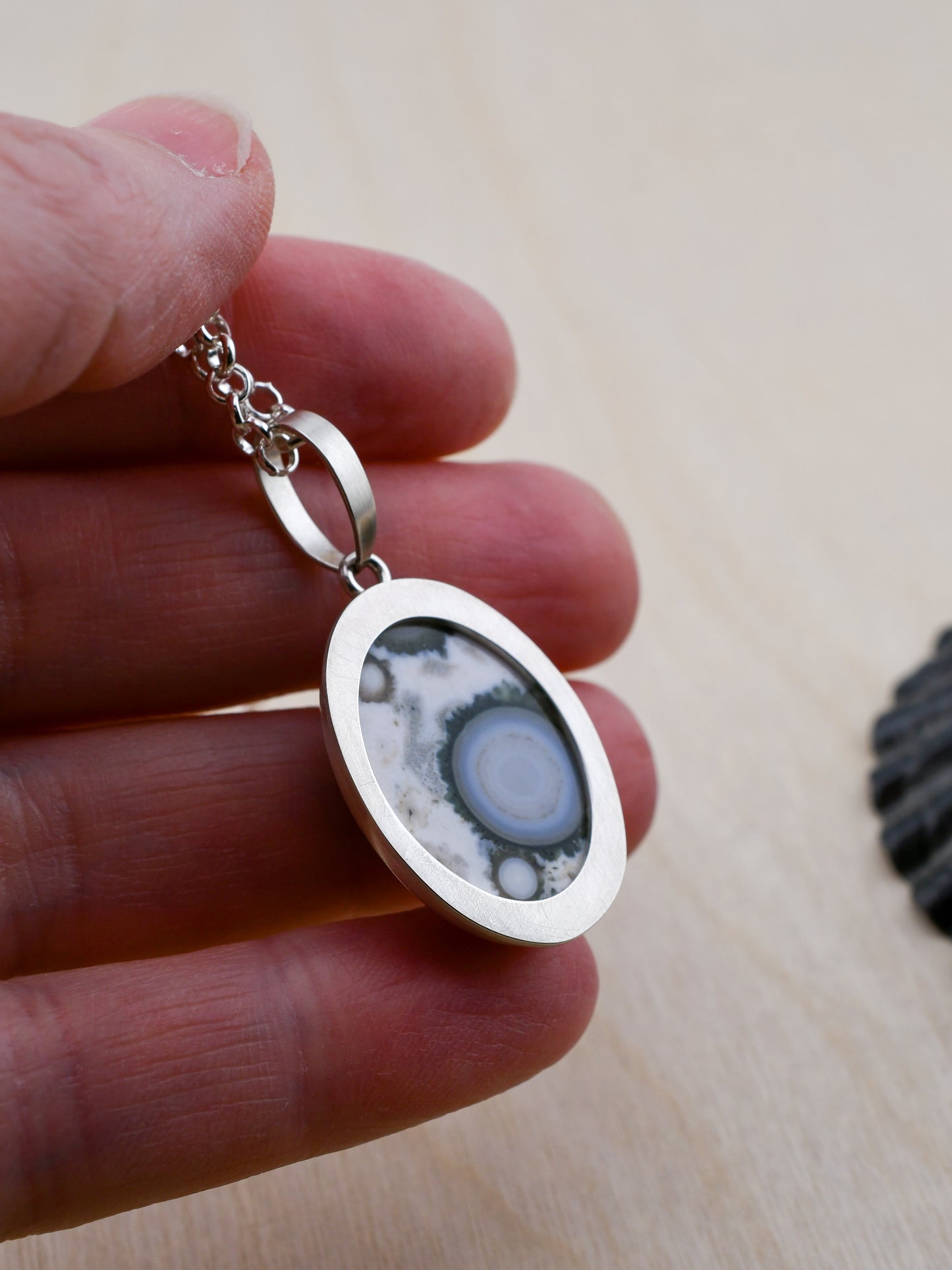 closeup of the backside of a silver pendant with a patterned stone held between fingers on a light wooden surface