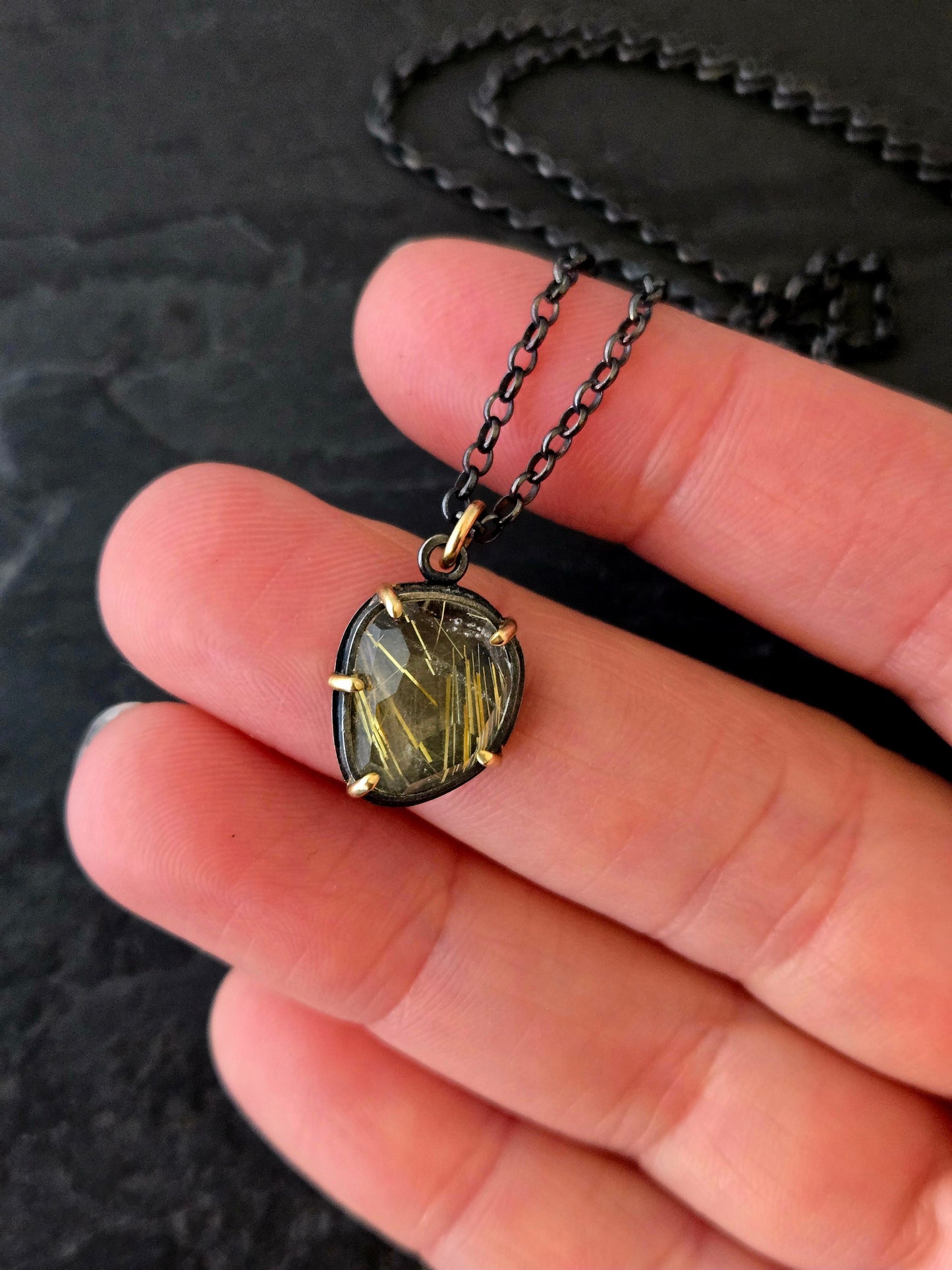 Rutilated quartz pendant set in 14k gold and black silver held in a hand over a dark textured surface