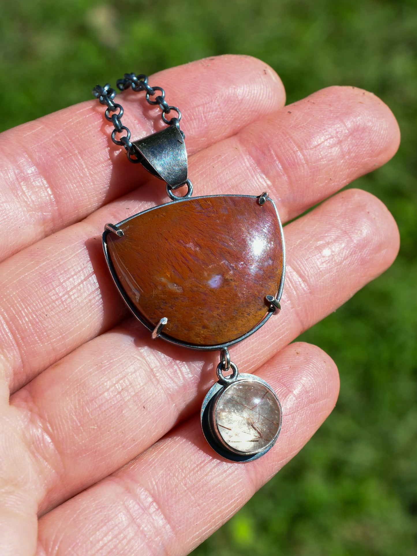 Hand holding a pendant with a large red stone and smaller round stone, against a blurred green background.