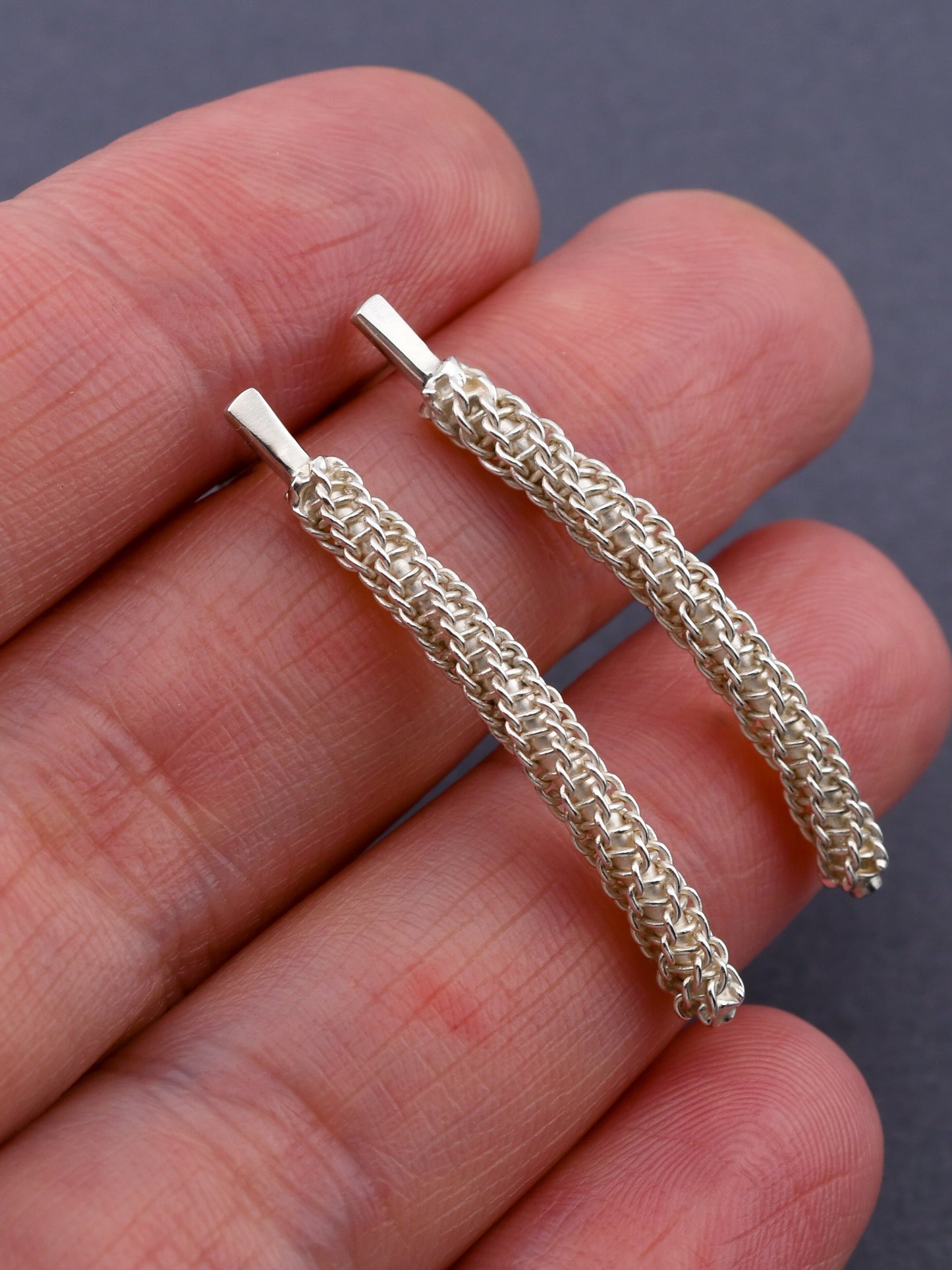 Two silver woven post earrings held between fingers against a dark background