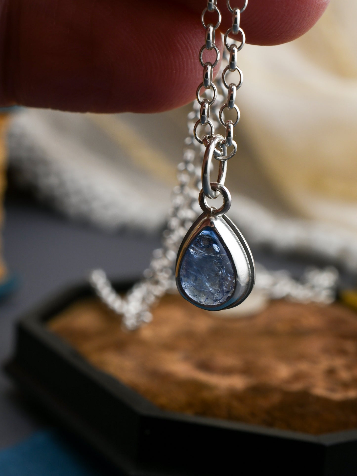 Teardrop-shaped blue pendant on a silver chain held by a hand.