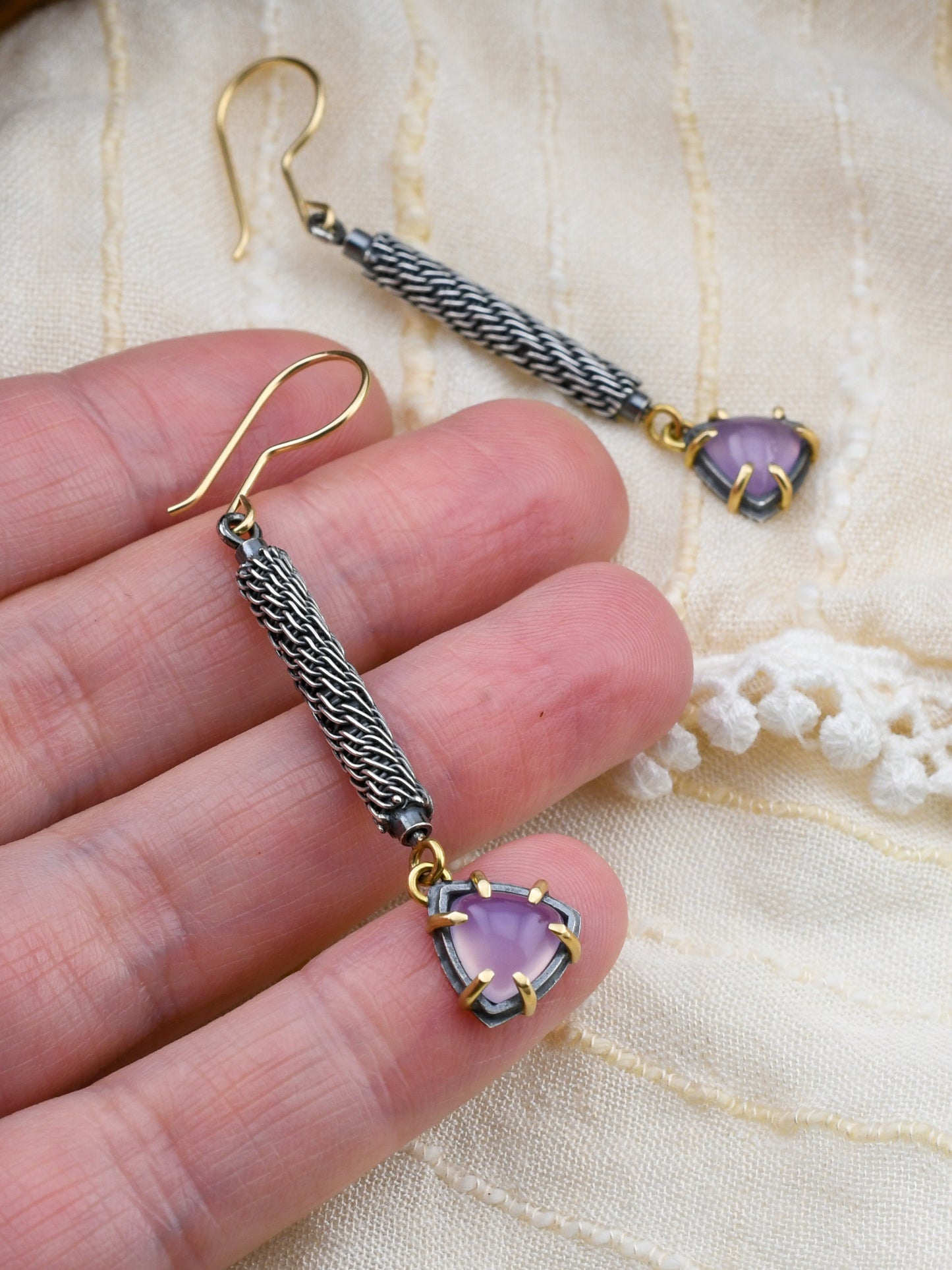 Gold and silver earrings with purple gemstones held by a hand on a textured fabric background