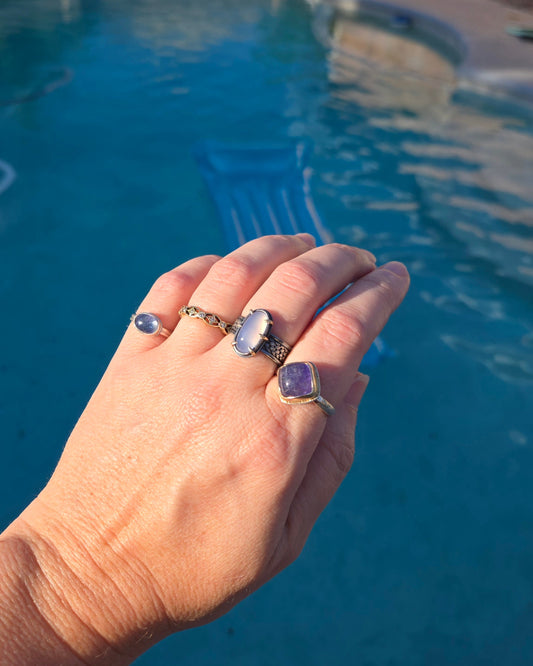 A hand poised over a blue swimming pool with four gemstone and diamond rings of silver and gold.