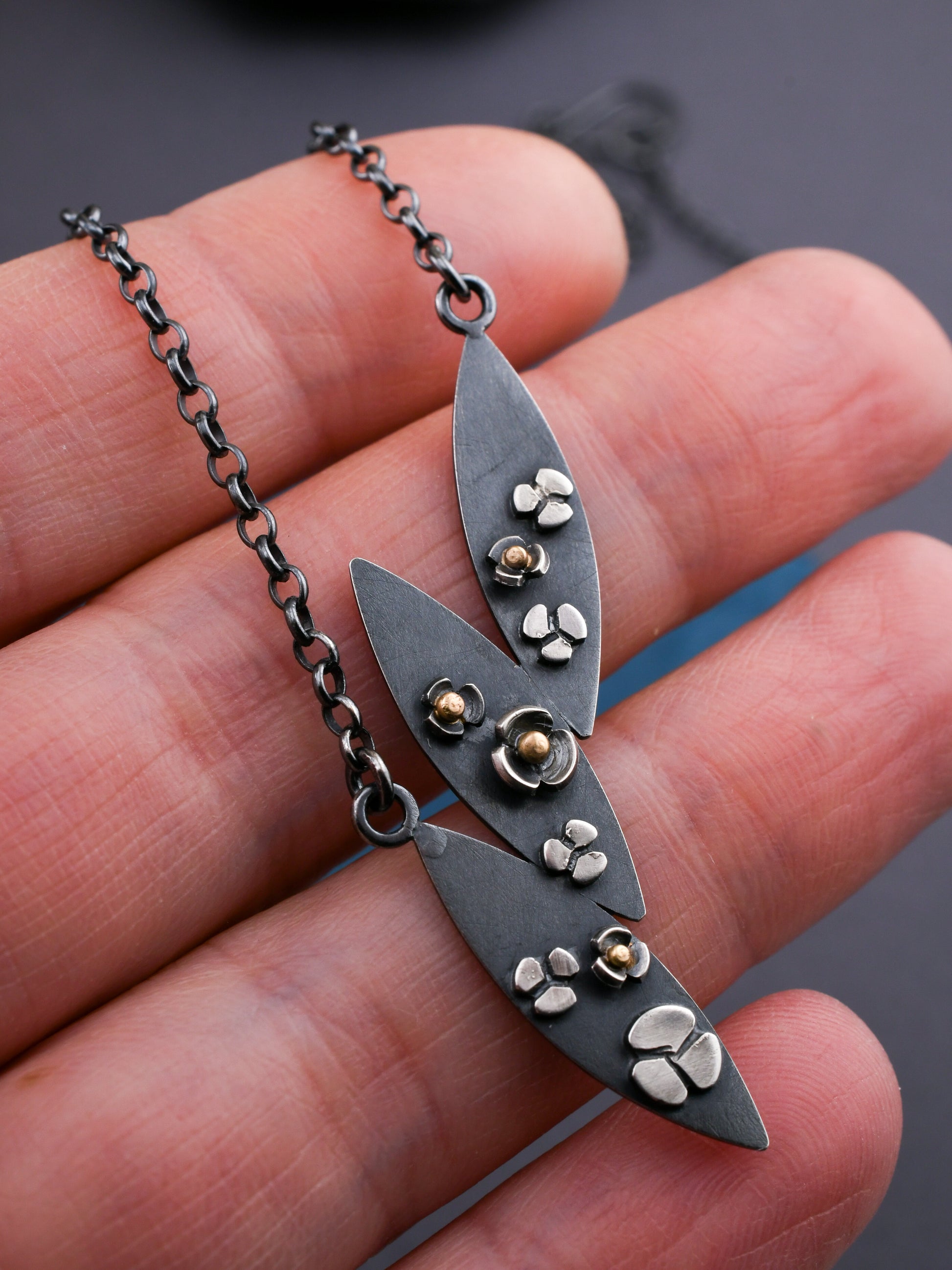 leaf shaped necklace with flowers and gold accents held in a hand with a dark gray background
