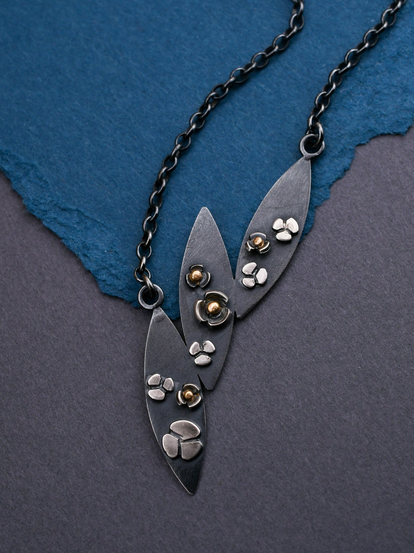 closeup of leaf necklace in black silver with gold accents on a textured blue and gray background