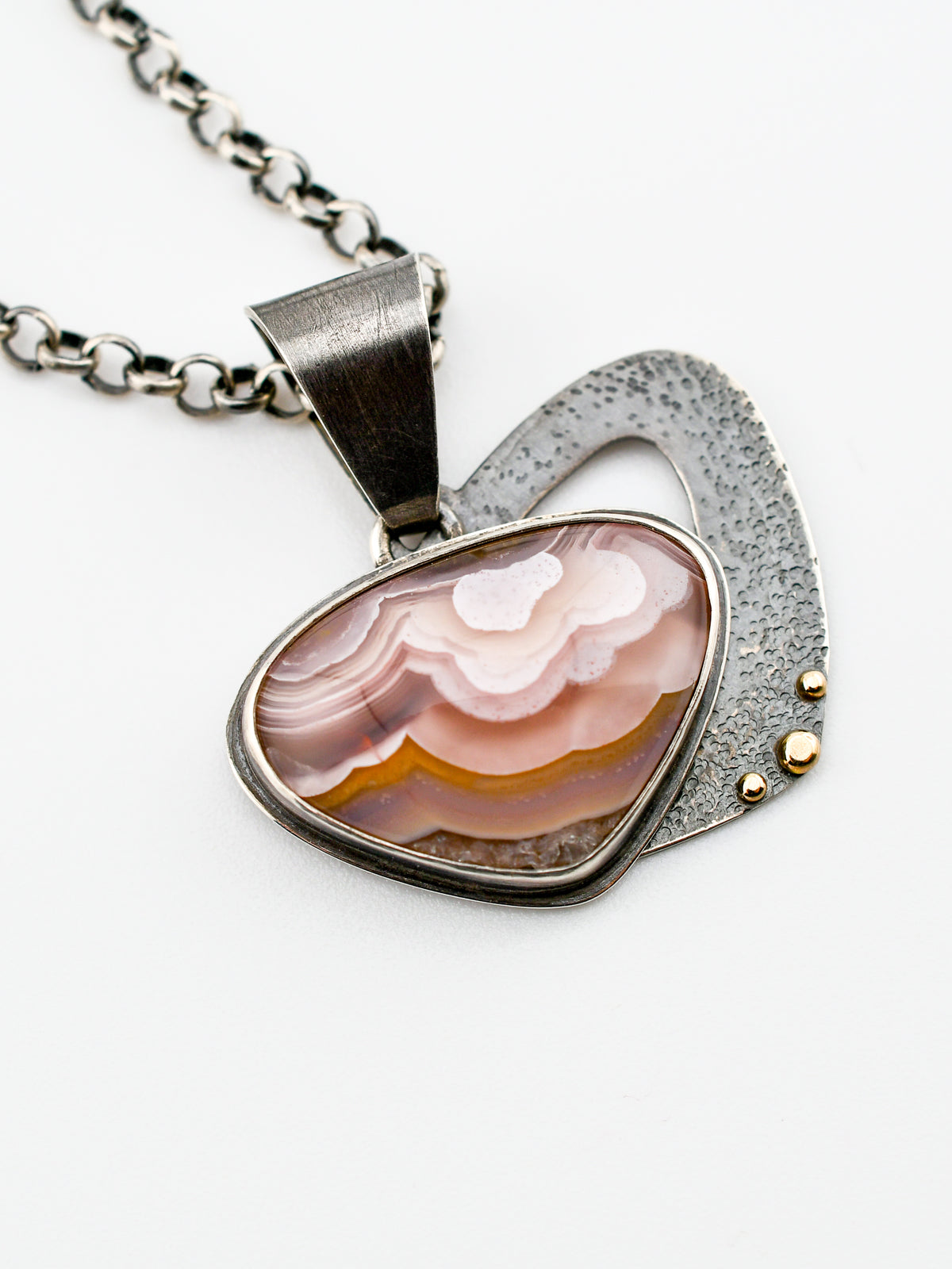 a rounded triangular Agua Nueva agate pendant with wavy patterns of pale pink and gold had a textured backplate that swings behind the stone and is accented with 14k gold granules, shown on a white background