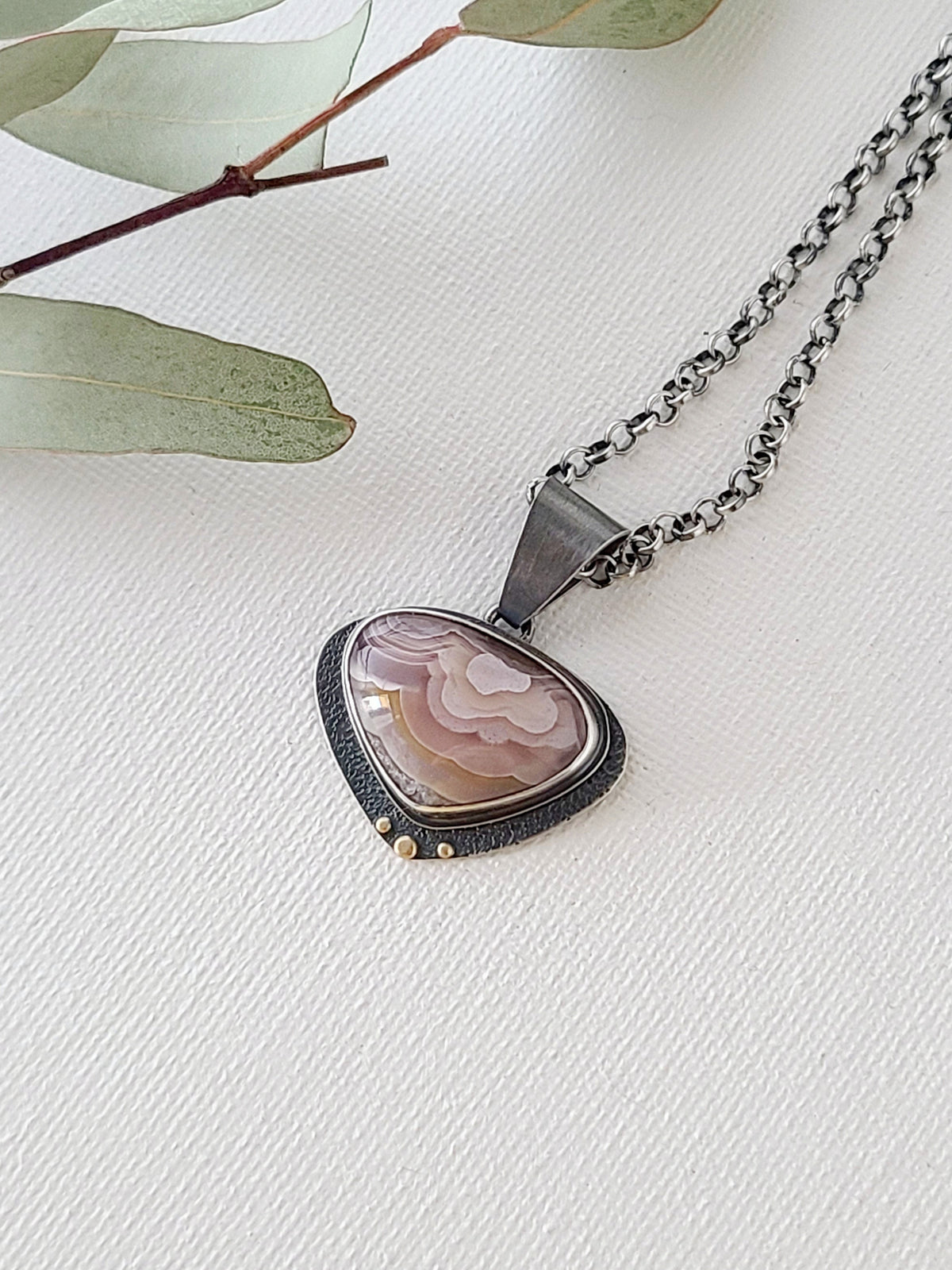 Pink and rose Agua Nueva agate pendant with an oxidized silver textured backplate adorned with 14k gold granules shown on a textured white background with pale green leaves