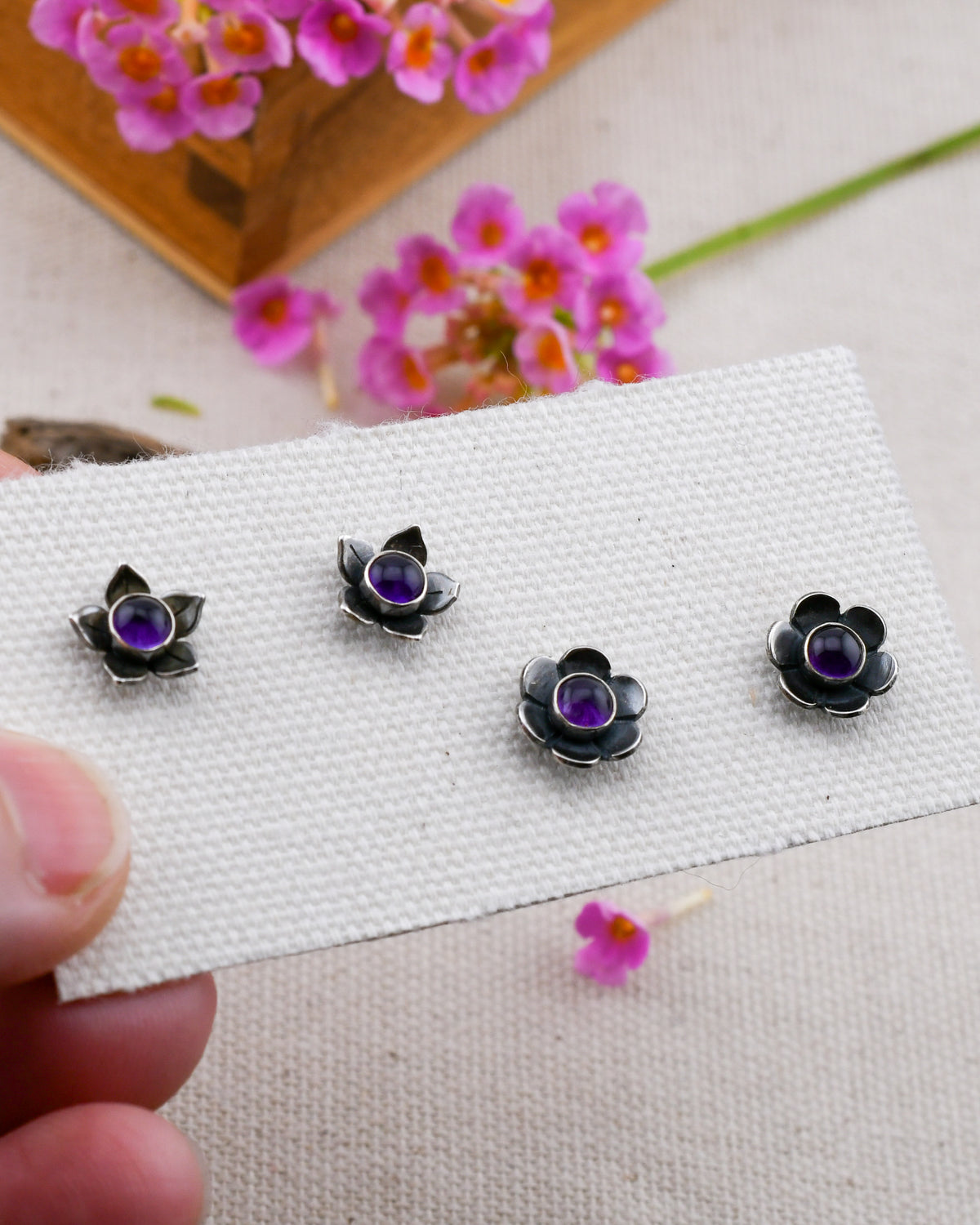two pairs of silver flower-shaped stud earrings with purple amethyst centers, displayed on a white textured card held by a hand against a light beige fabric background with pink flowers.