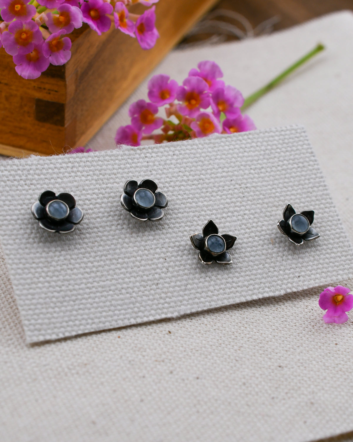 Two pairs of small black flower-shaped stud earrings with round blue aquamarine centers, arranged on a textured white fabric surface with pink flowers and a wooden box in the background.