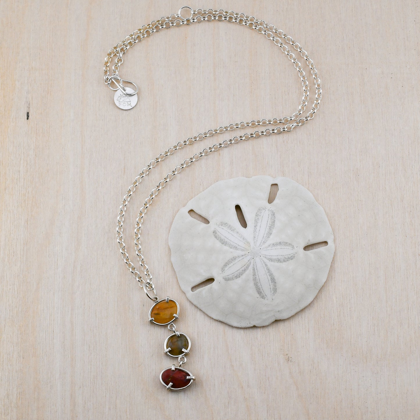 yellow green and red beach stone pendant on a silver chain displayed next to a sand dollar on a pale wood background