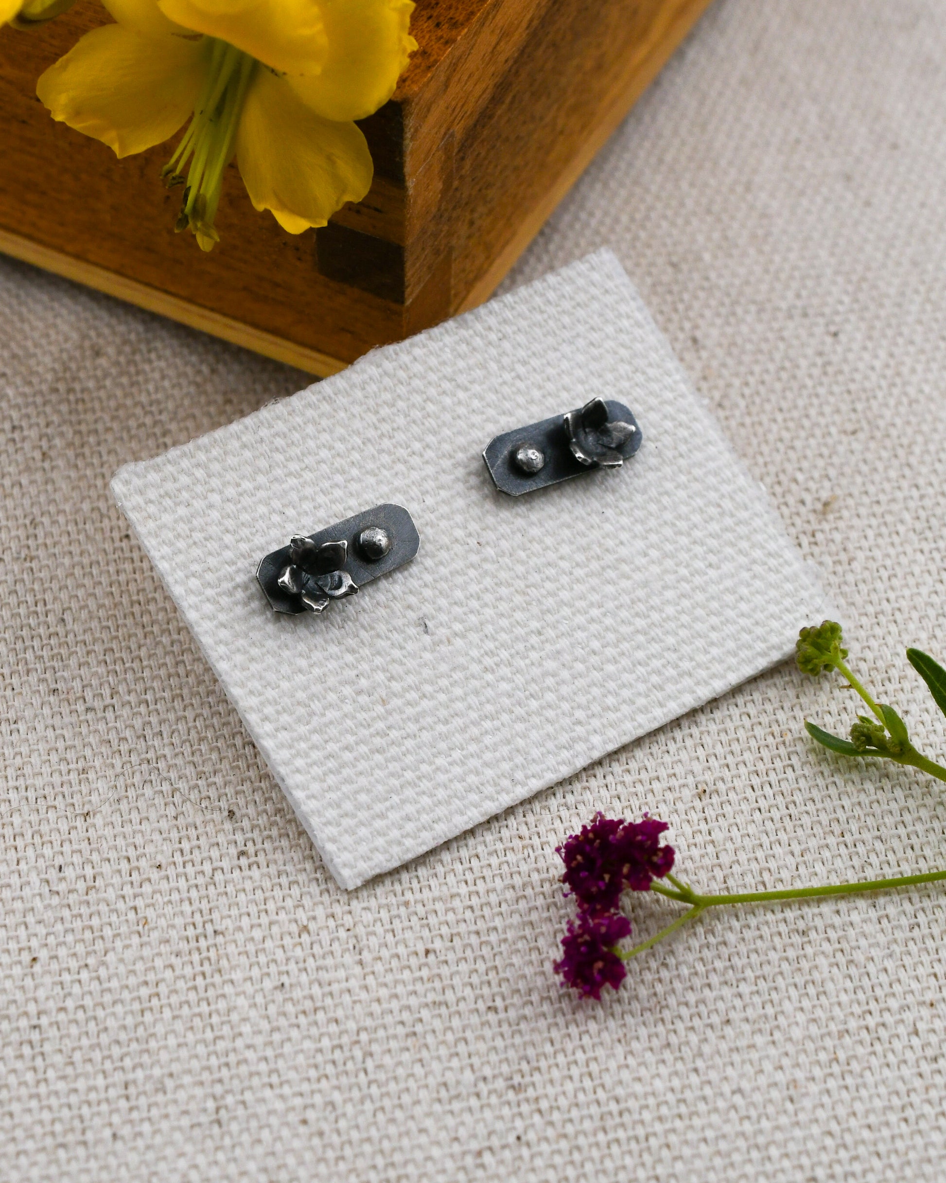 A pair of blackened silver stud earrings with flowers and a silver granule on a bar-shaped backing displayed on a small white textured card. The card is placed on a beige linen surface with a yellow flower in a wooden box.