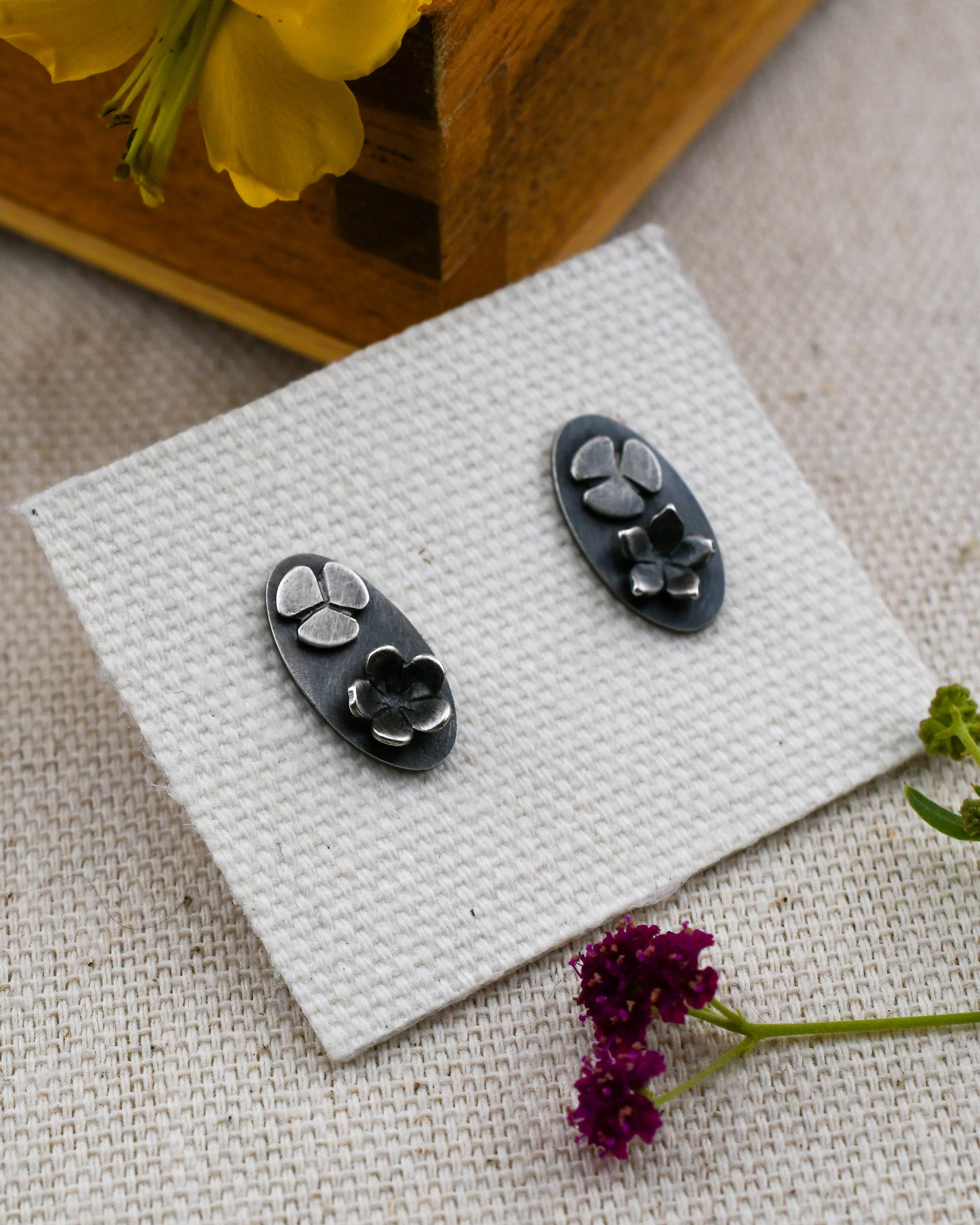 A pair of blackened silver oval stud earrings with two silver flowers blooming on the surface displayed on a textured white card with small flowers and a wooden box in the background.