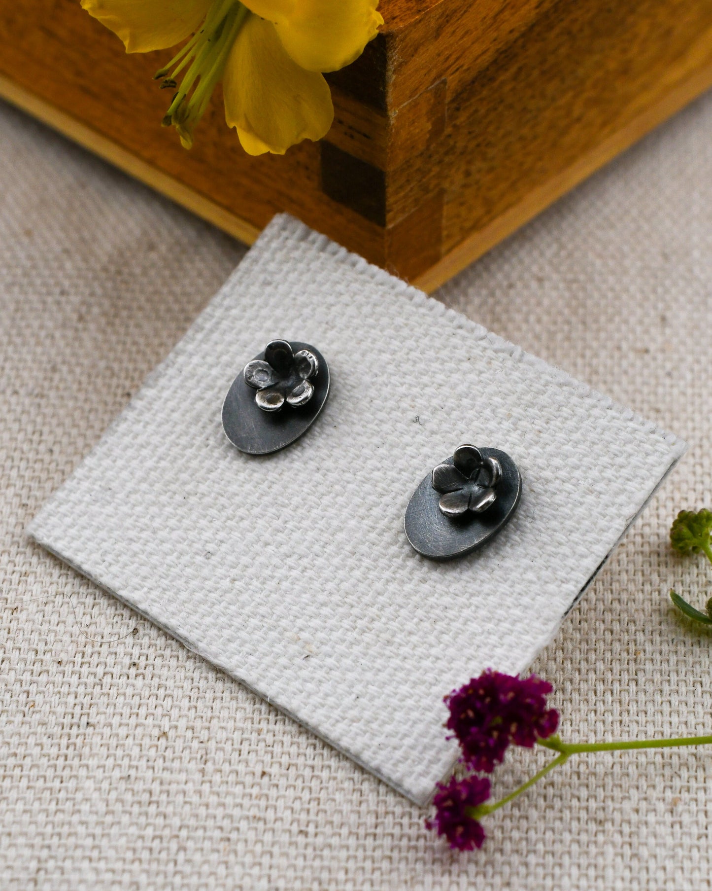 A pair of small blackened silver stud earrings with flowers on a small oval backing displayed on a small white card, placed on a textured beige fabric with a wooden box edge and small flowers nearby.