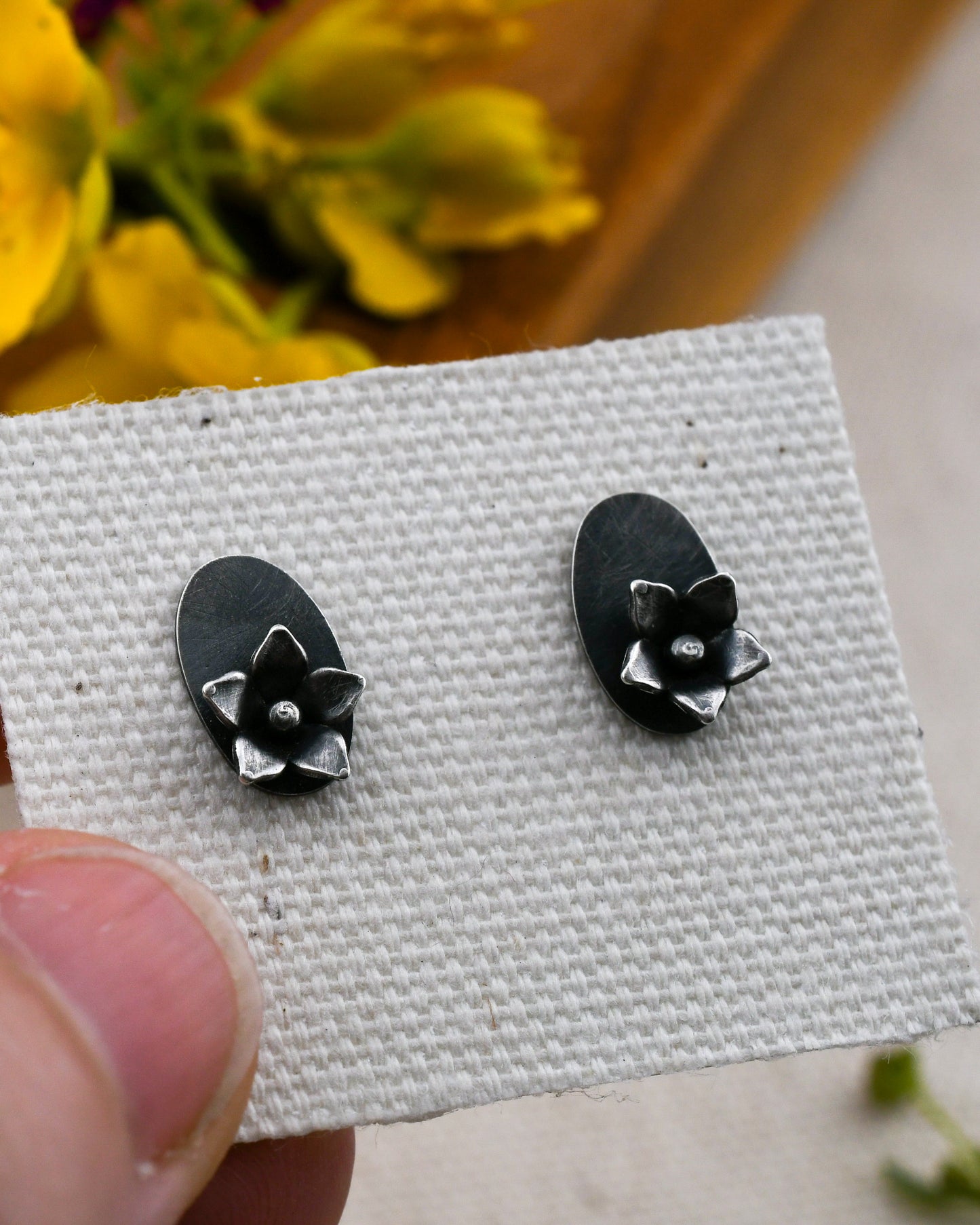 A pair of blackened silver stud earrings with flower on an oval backing displayed on a textured white card, held by a person's thumb with blurred yellow flowers in the background.