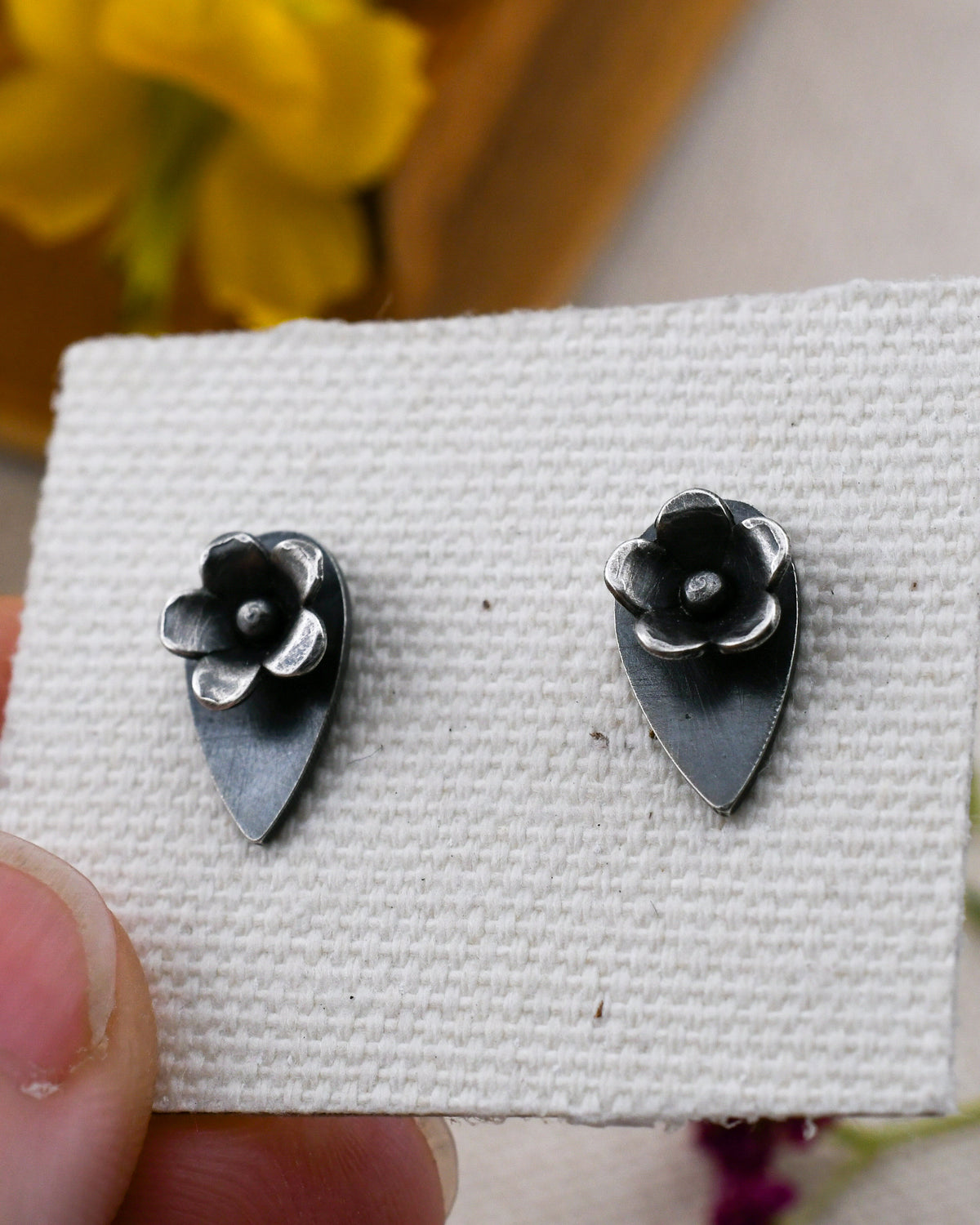 A pair of small  silver stud earrings  with flowers on talon-shaped backing, displayed on a textured white card held by a hand.