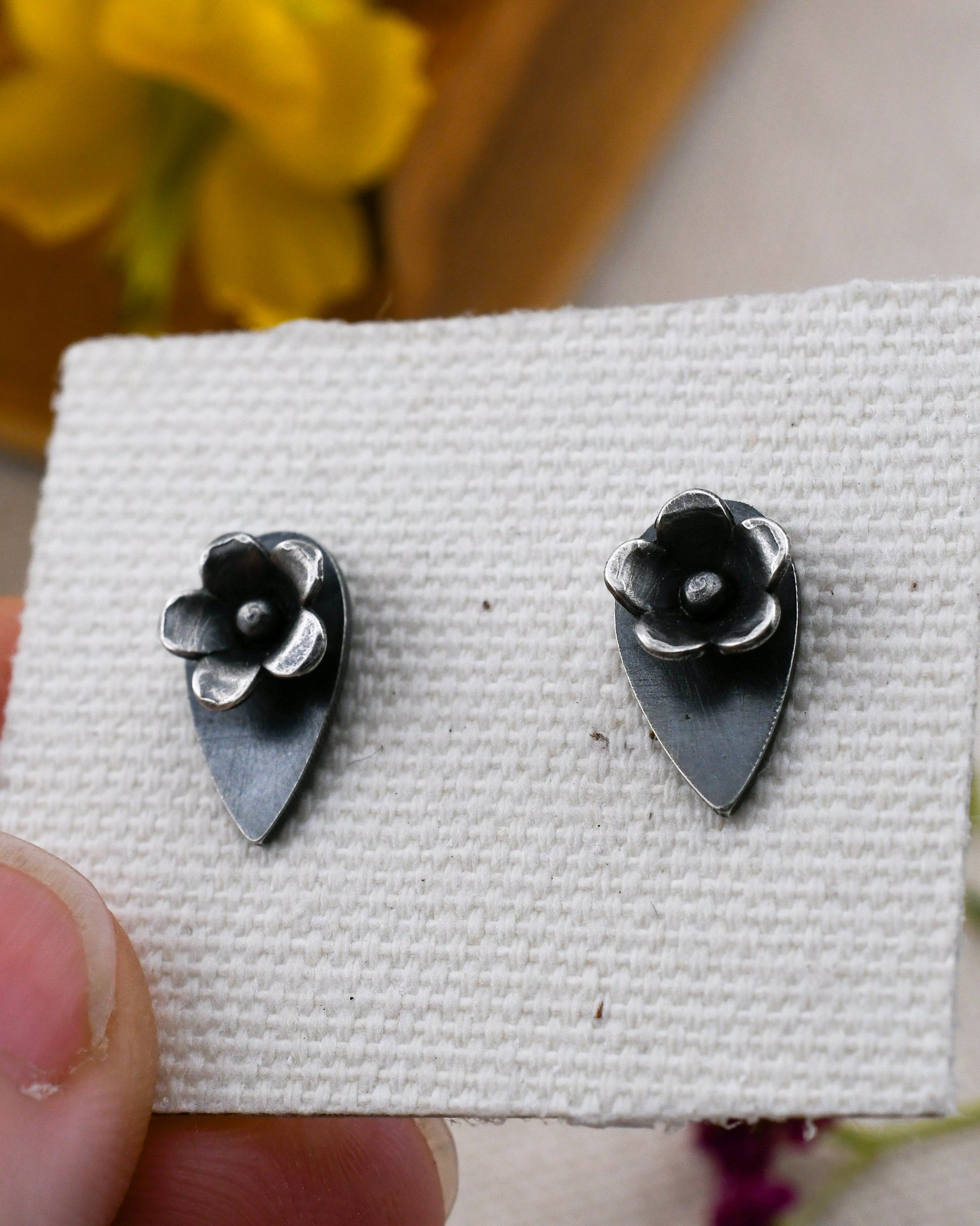 A pair of small  silver stud earrings  with flowers on talon-shaped backing, displayed on a textured white card held by a hand.