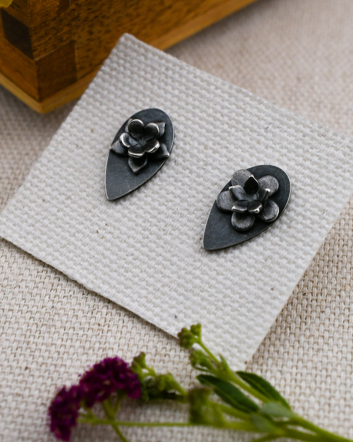 A pair of blackened silver flower stud earrings displayed on a textured white card, with a wooden box corner and pink flowers in the background.
