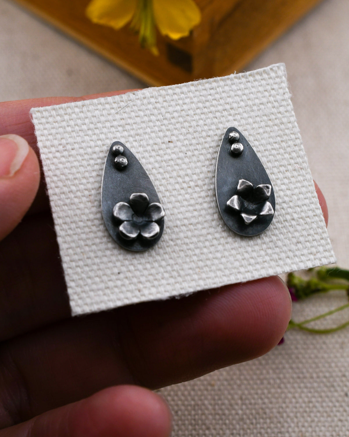 A pair of blackened silver floral teardrop stud earrings displayed on a textured white card held by a hand.
