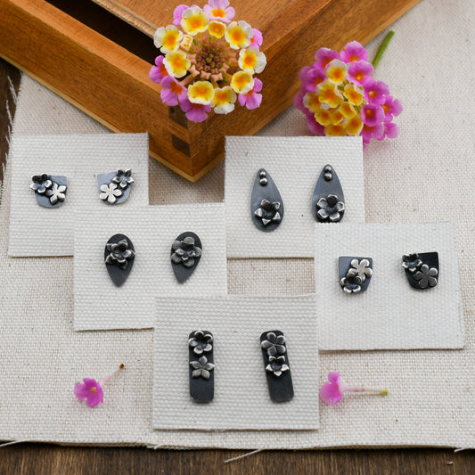 5 pairs of blackened silver stud earrings with flower details displayed on linen cards on a beige and wood background with pink and yellow flowers.