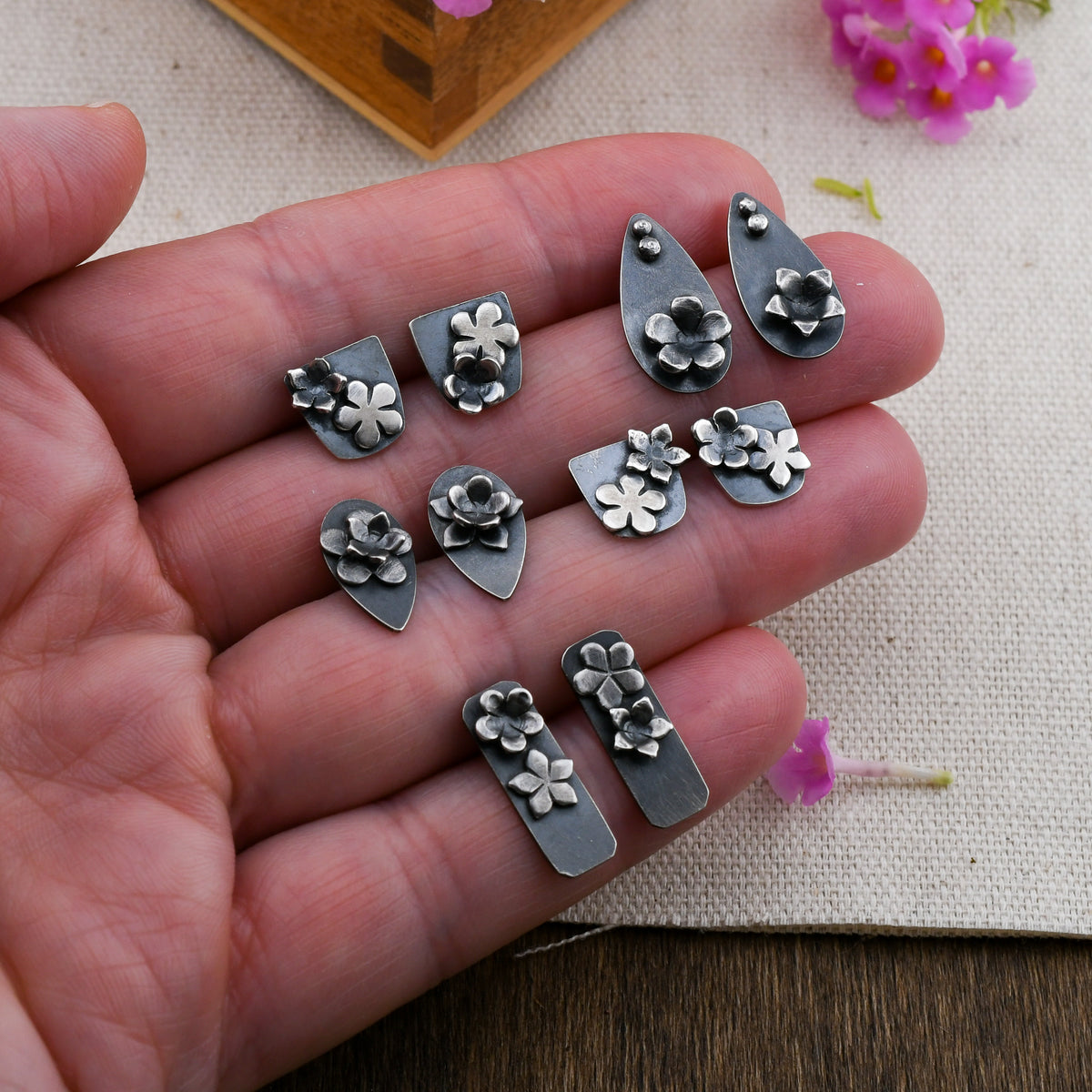 A hand holding 5 pairs of blackened silver stud earrings with floral details over a beige background with pink flowers and wood