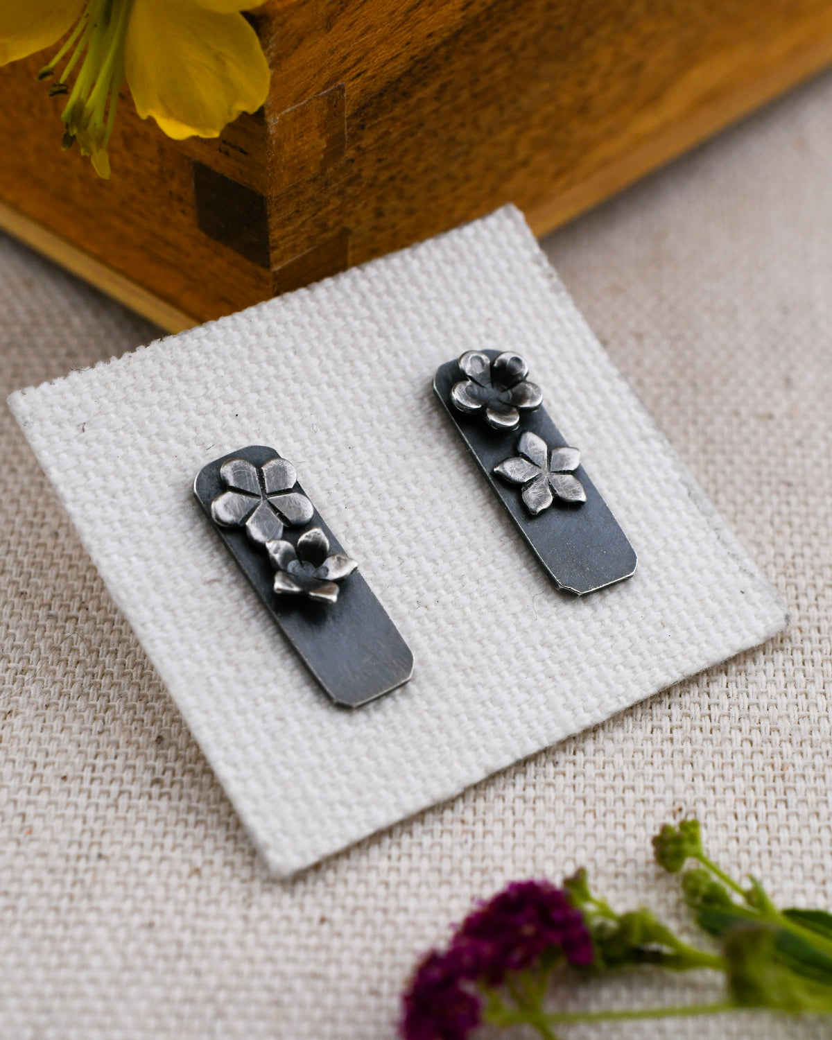 A pair of blackened silver rectangular stud earrings with raised silver flower details, displayed on a small white textured card over a woven fabric surface with a wooden box and dried flowers in the background.