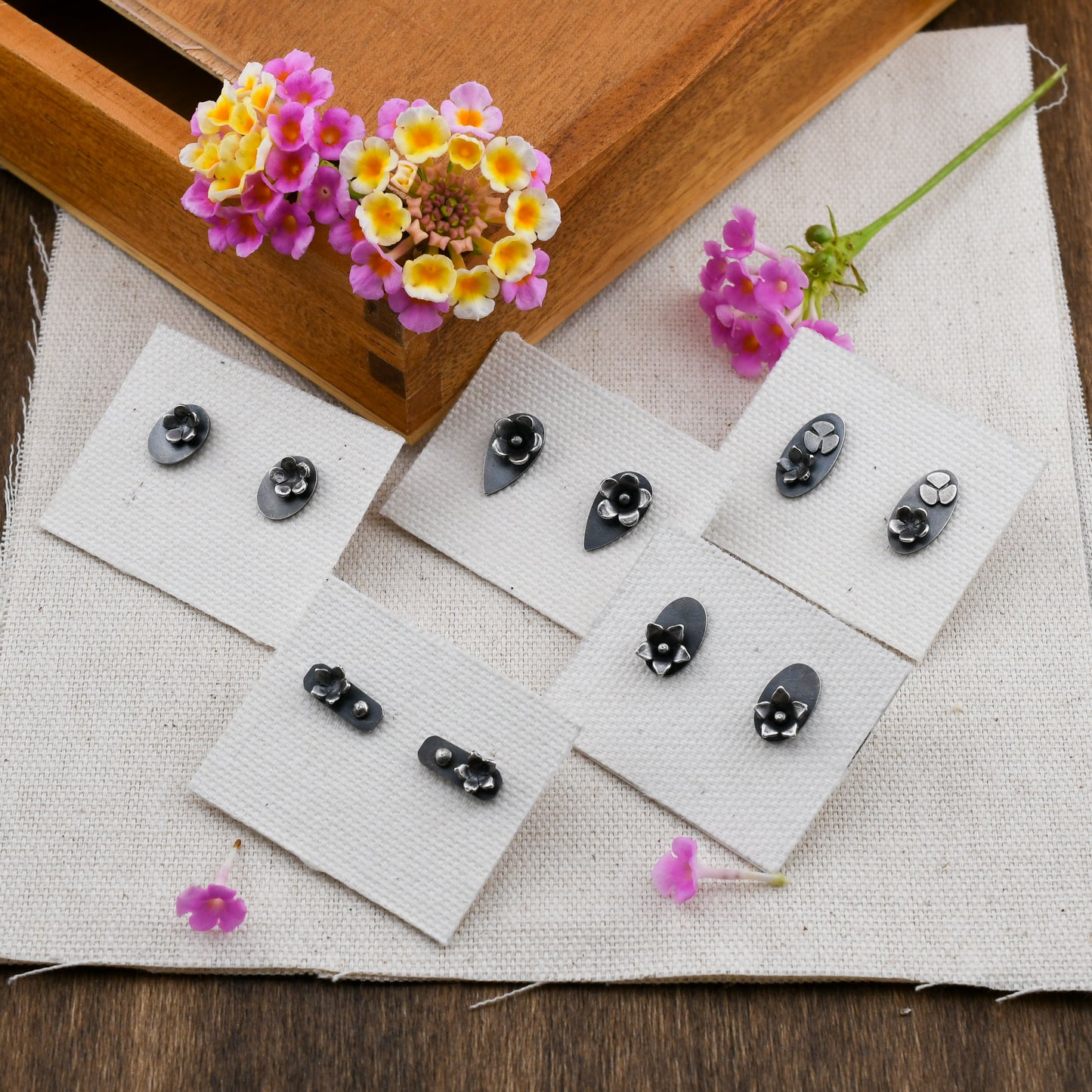 5 pairs of blackened silver stud earrings with flower details displayed on linen cards on a beige background with pink and yellow flowers in a wooden box