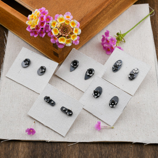 5 pairs of blackened silver stud earrings with flower details displayed on linen cards on a beige background with pink and yellow flowers in a wooden box
