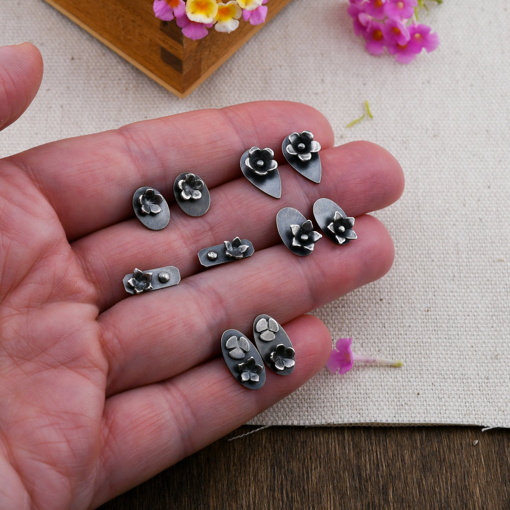 A hand holding 5 pairs of stud earrings with flower details over a beige background with pink and yellow flower and wood.