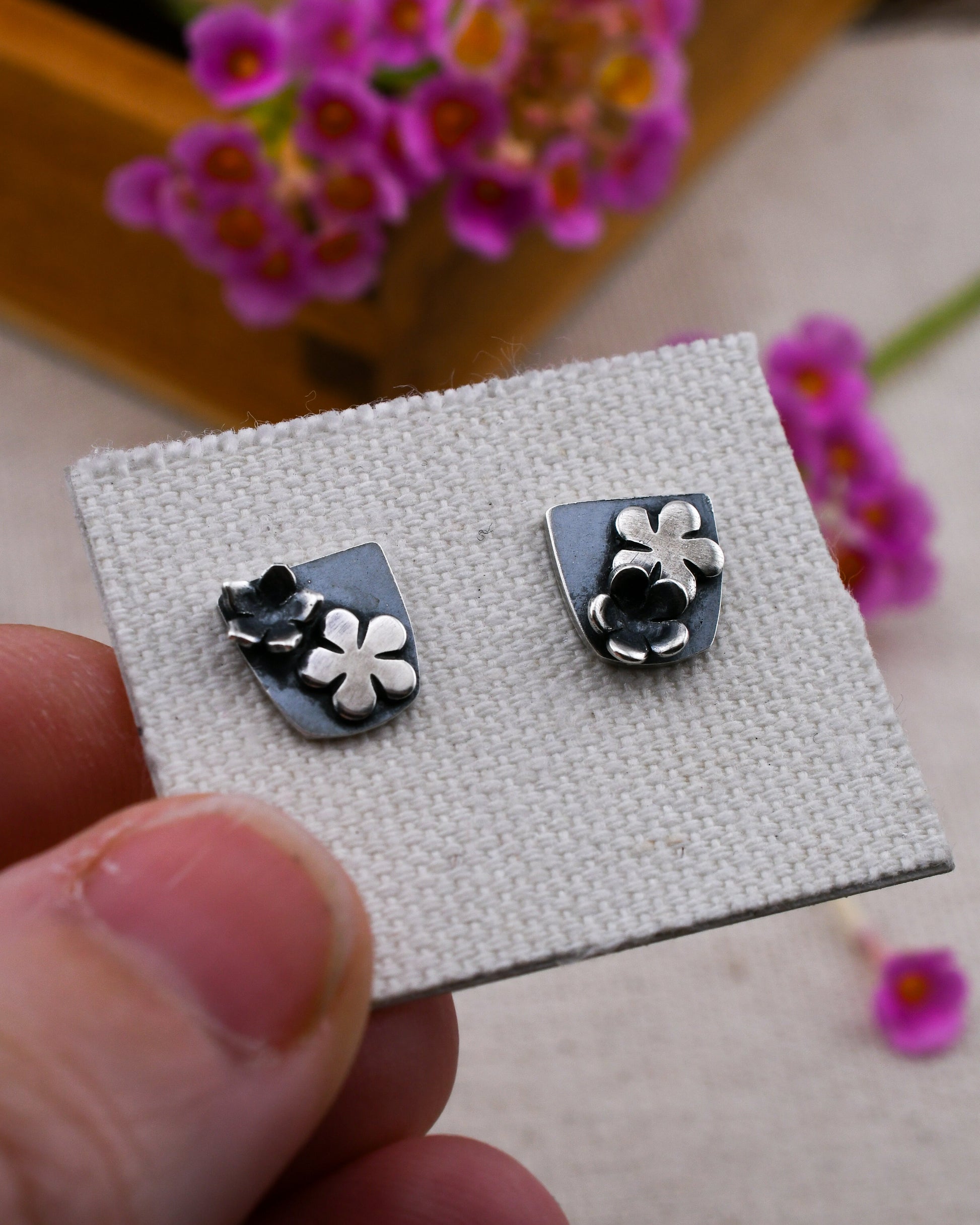 A close-up of a hand holding a textured display card with a pair of silver stud earrings. Each earring features a shield-shaped silver back with small silver flowers, set against a soft-focus background of pink flowers.