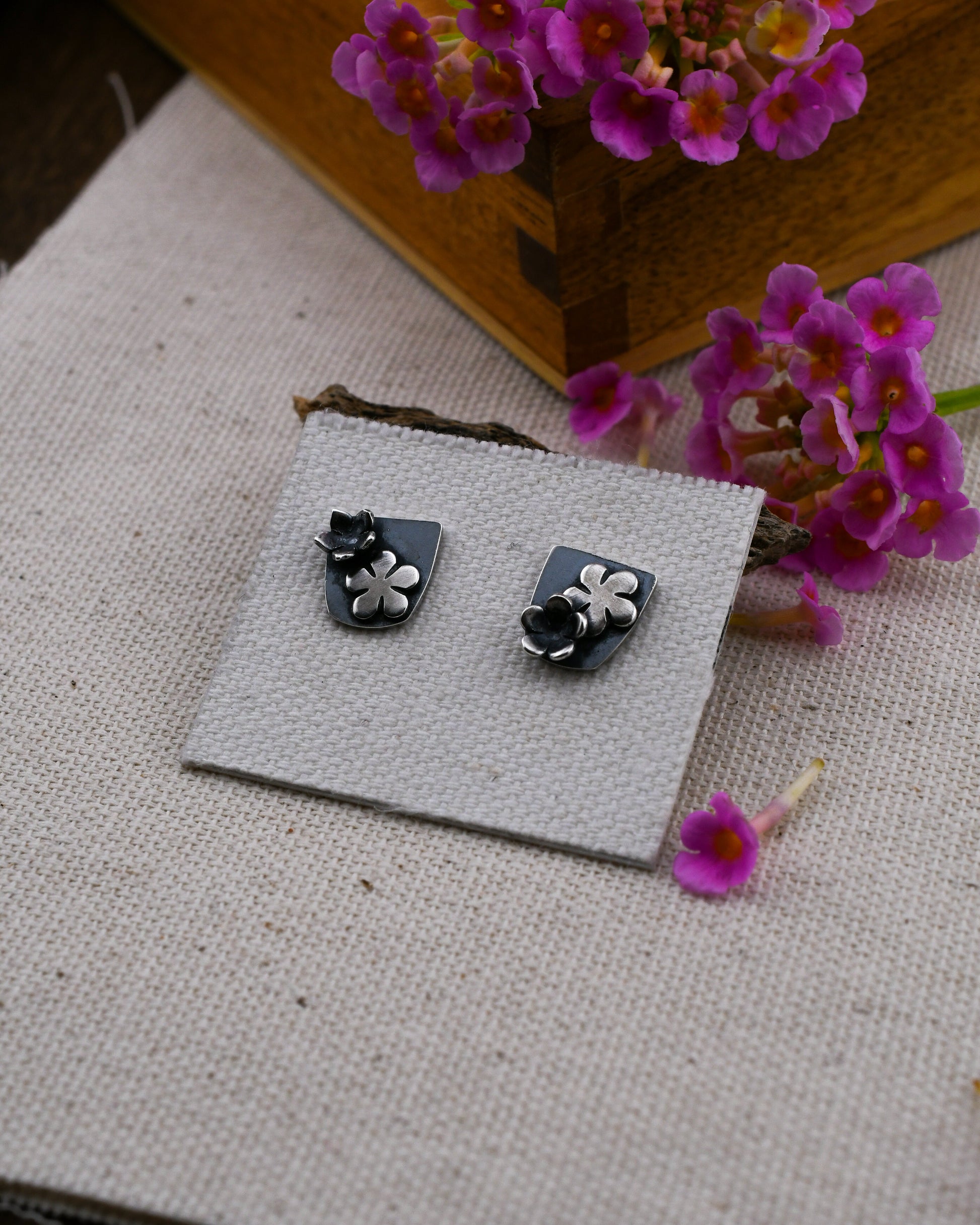 A pair of black and silver flower stud earrings displayed on a small square card, set on a textured beige fabric with pink flowers and a wooden box in the background.