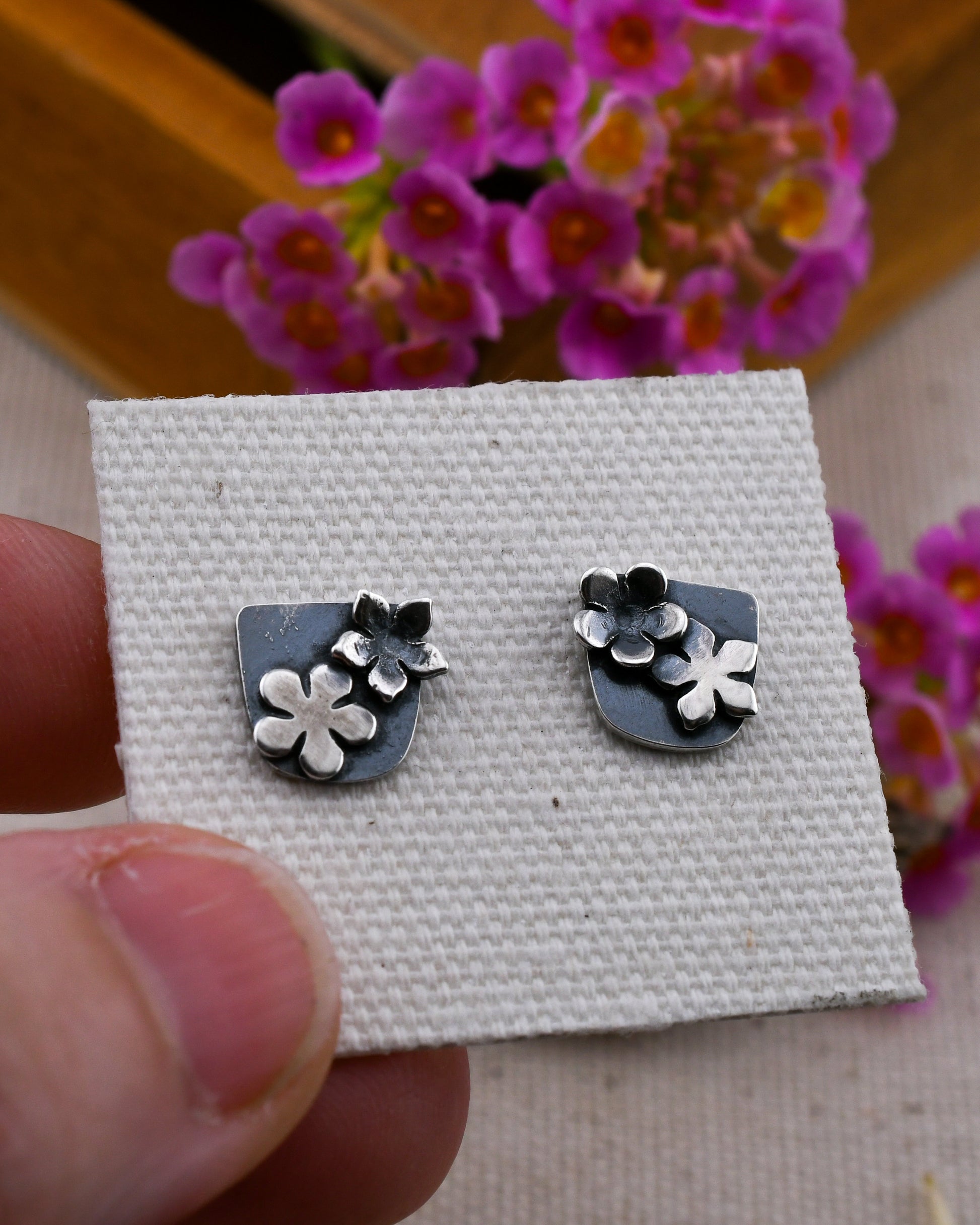 A pair of silver stud earrings with small flower and shield motifs, displayed on a textured white card held by a hand, with pink flowers blurred in the background.