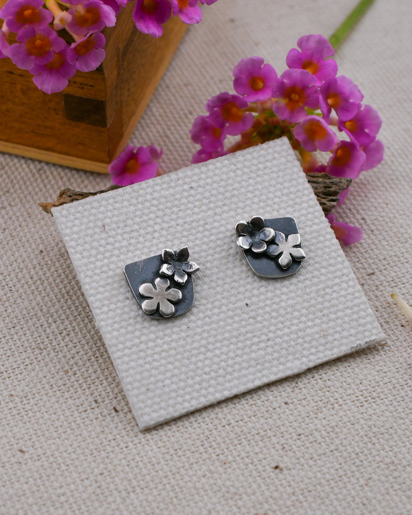 A pair of silver flower stud earrings displayed on a textured white card, set against a beige linen background with pink flowers in the corner.