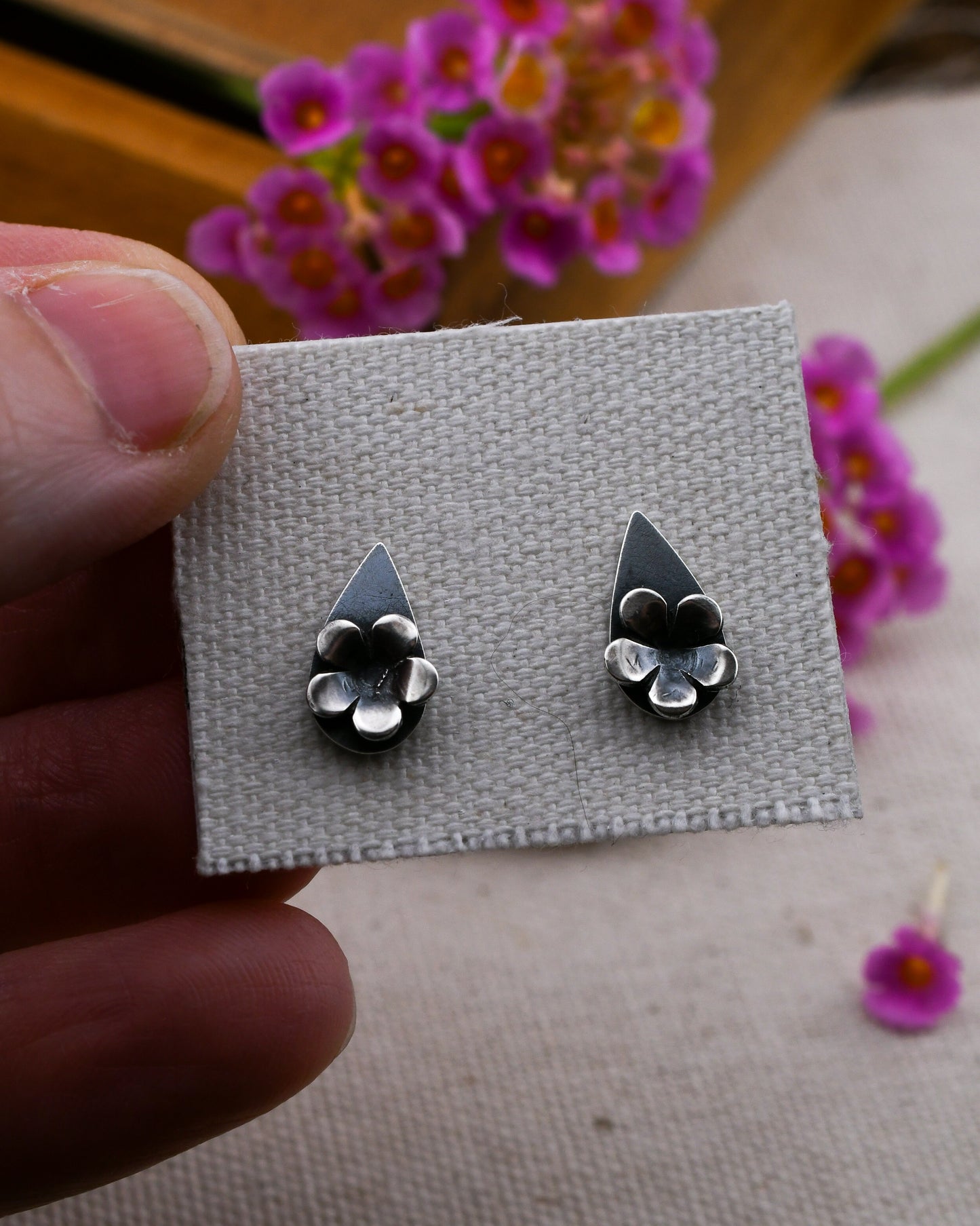 A close-up of a hand holding a small gray fabric card displaying a pair of teardrop silver stud earrings with silver blossoms. The background is softly blurred with pink flowers and a wooden surface.
