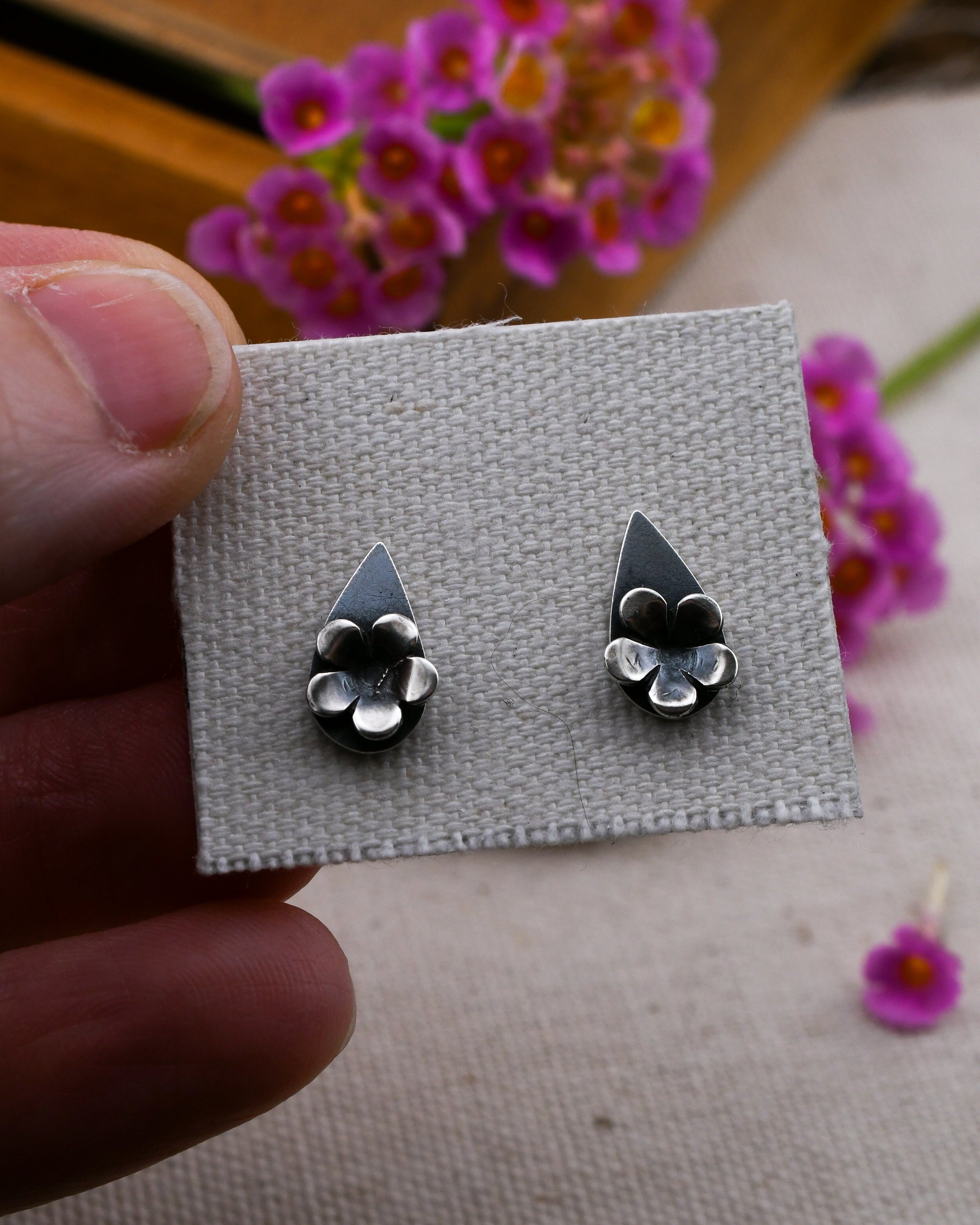 A close-up of a hand holding a small gray fabric card displaying a pair of teardrop silver stud earrings with silver blossoms. The background is softly blurred with pink flowers and a wooden surface.