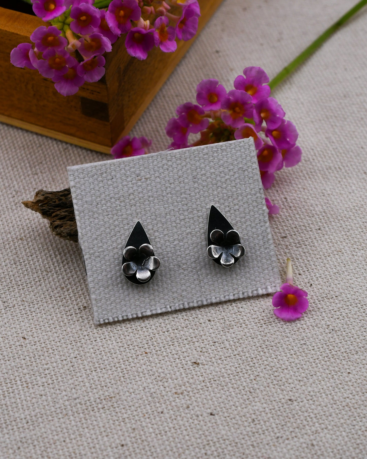 A pair of black and silver teardrop shaped floral stud earrings displayed on a small gray card, set against a textured beige fabric with pink flowers and a wooden box in the background.