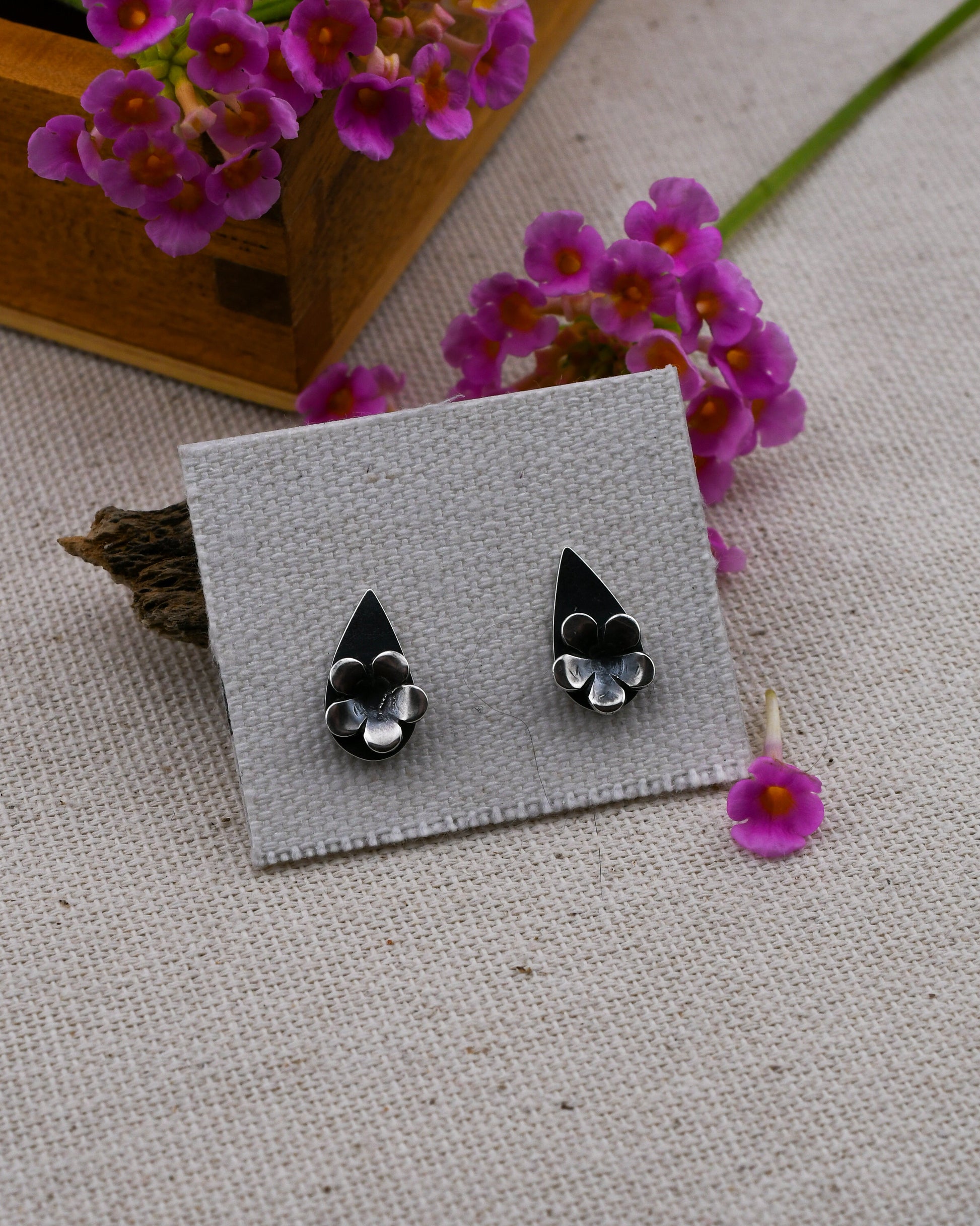 A pair of black and silver teardrop shaped floral stud earrings displayed on a small gray card, set against a textured beige fabric with pink flowers and a wooden box in the background.