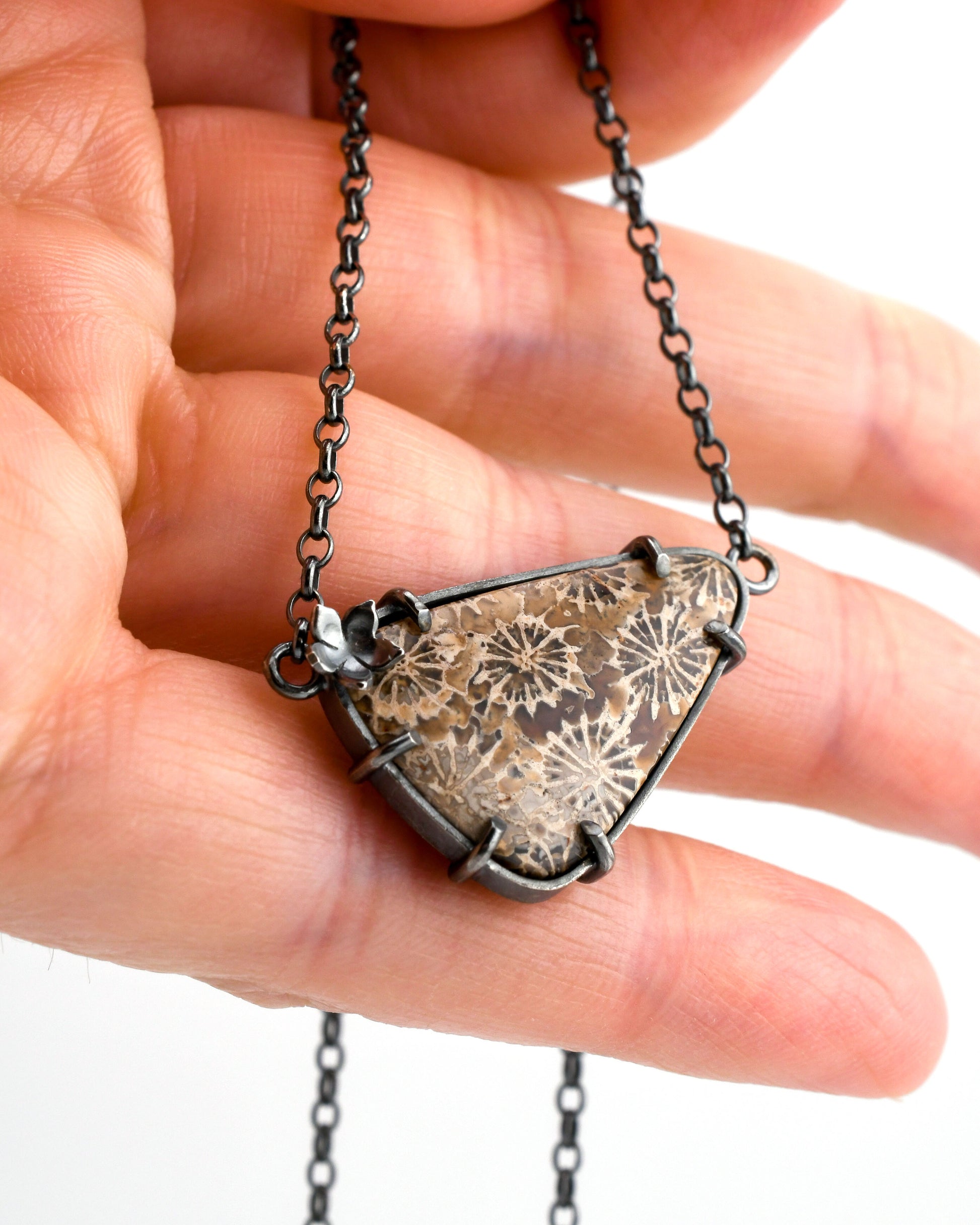Triangular fossil coral pendant with earth tone floral patterns and silver flower accents held in a hand on a white background