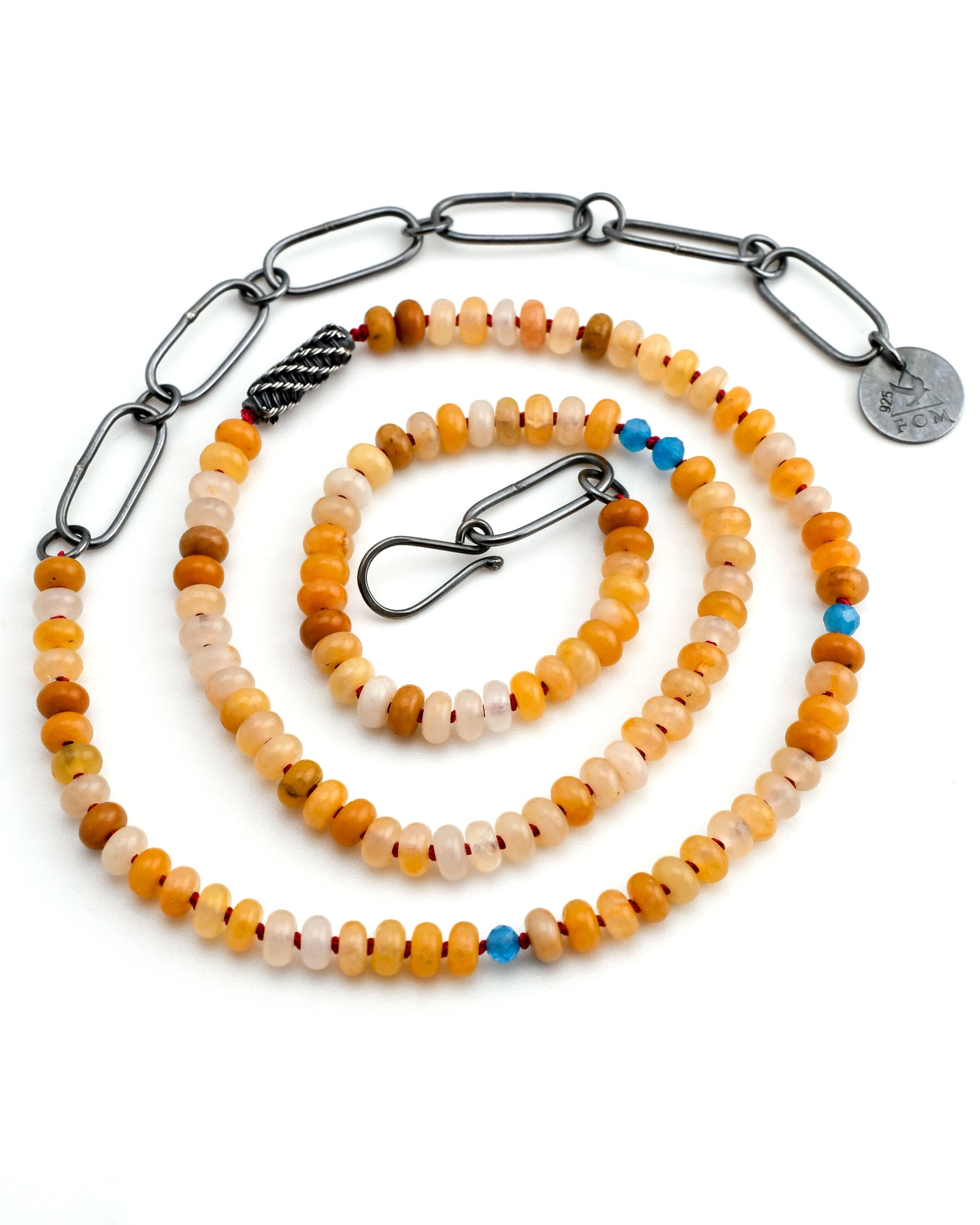 yellow jade and blue apatite beaded necklace on red silk with woven silver bead and black silver chain arranged in a spiral on a white background