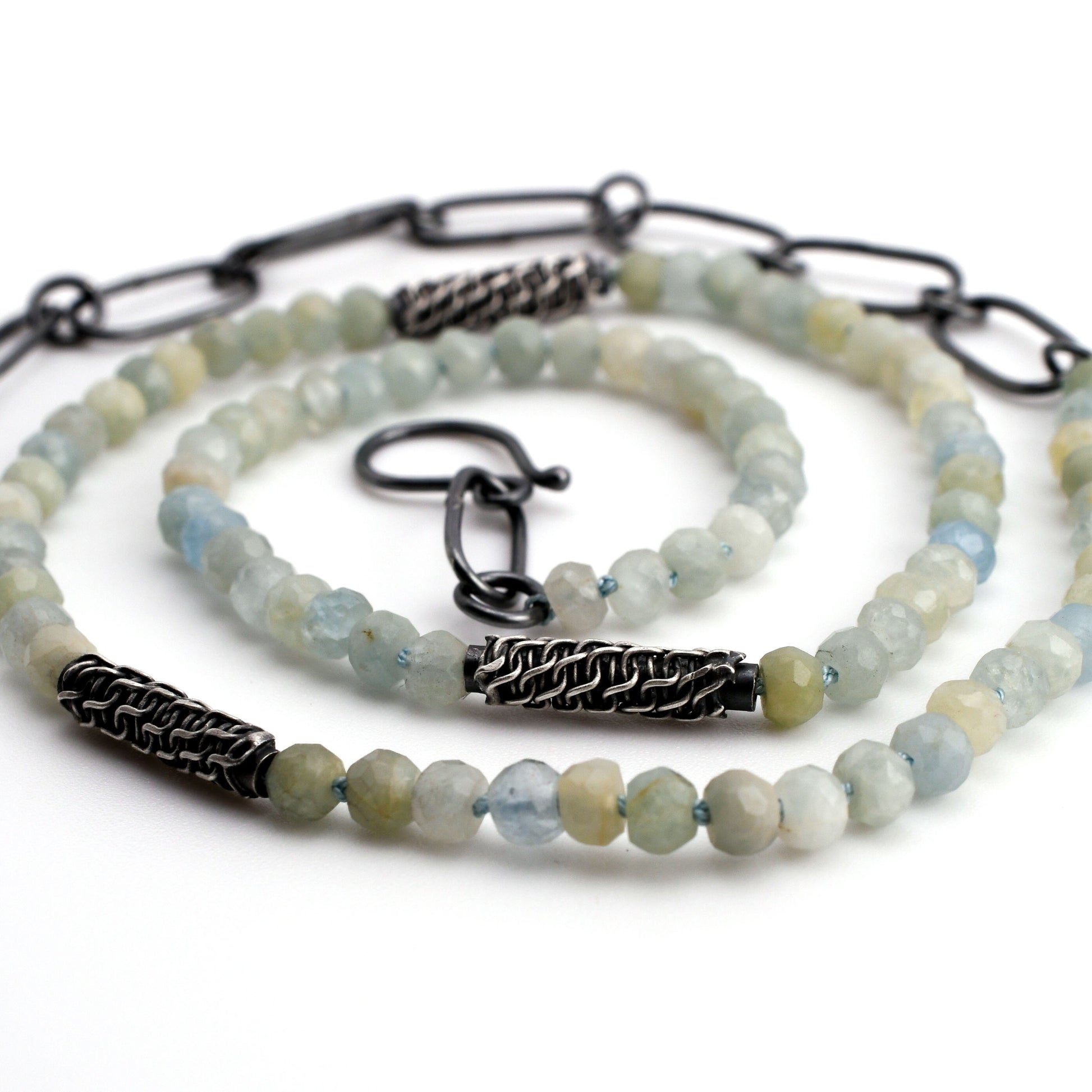 faceted natural blue aquamarine beaded necklace with woven silver beads and black silver chain extender arranged in a spiral on a  white background