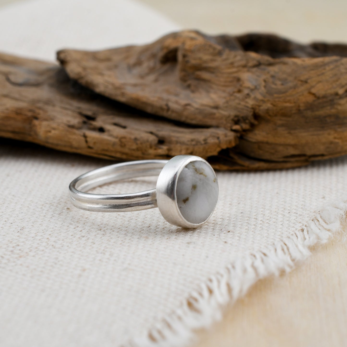 Silver ring with a round white beach stone on a textured fabric surface with driftwood in the background