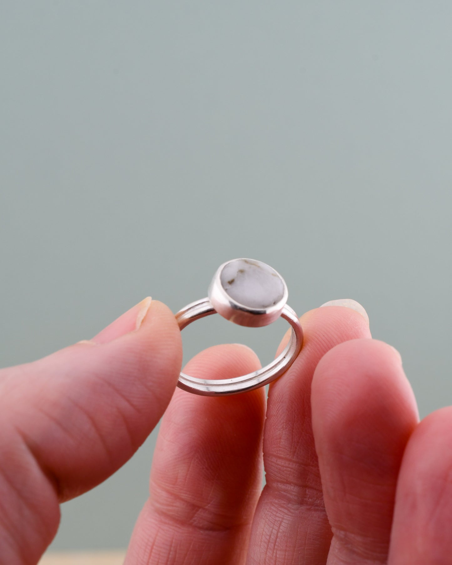 Silver ring with white stone held between fingers against a neutral background